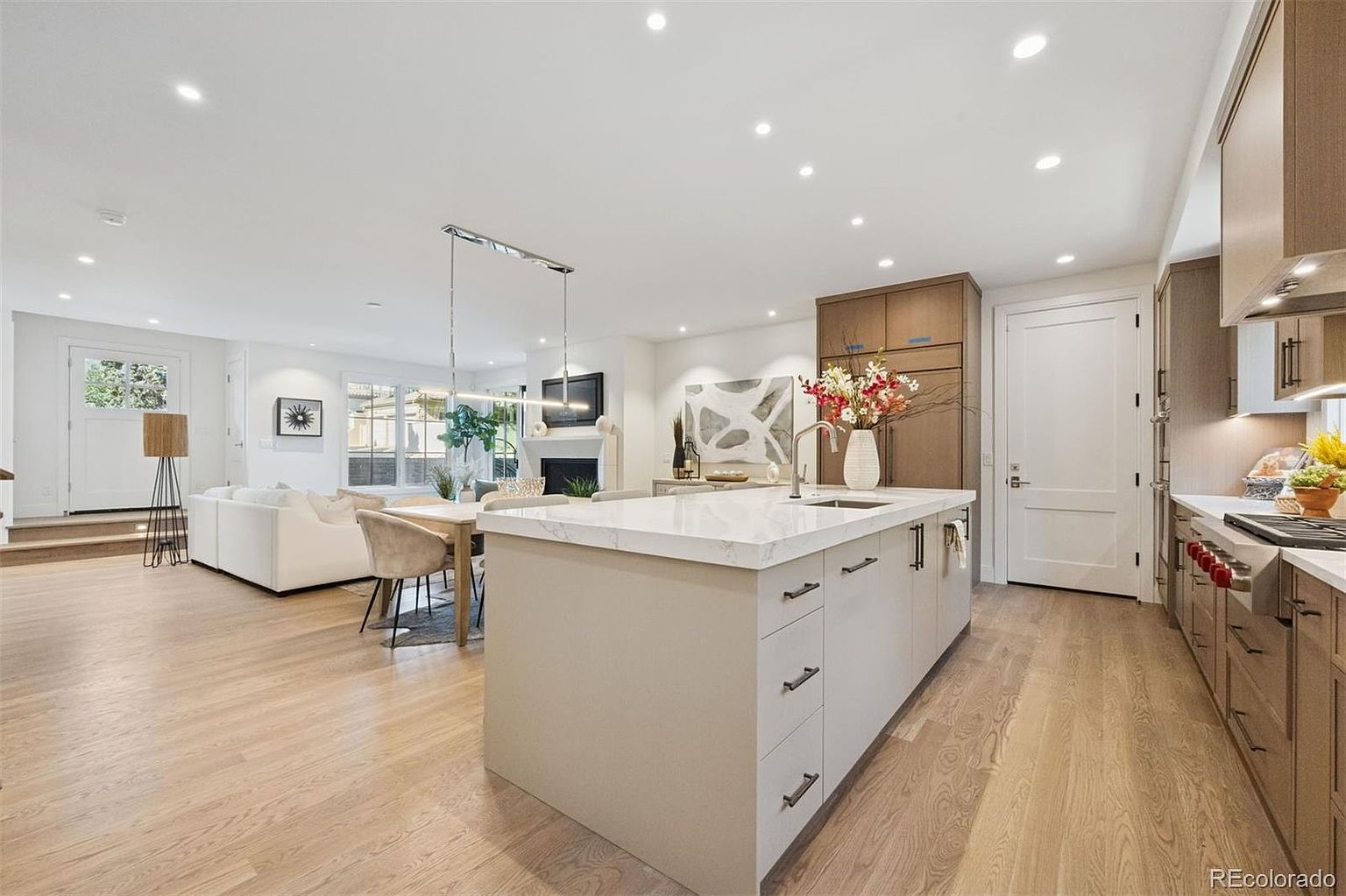This is a bright and open kitchen and living area. The kitchen features a large island with a white countertop and light beige cabinetry, stainless steel appliances, and a modern design. The living area includes a white sofa, a dining table with chairs, and a fireplace, creating a seamless and inviting space.