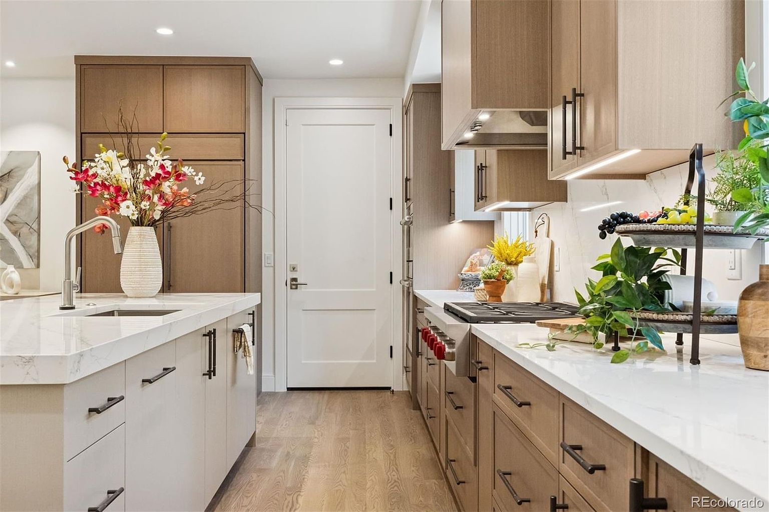 This is a well-lit kitchen featuring a modern design with light wood and white cabinetry, complemented by dark hardware. The countertops are a bright white marble, and the space is decorated with plants and flowers, adding a touch of nature. Stainless steel appliances and a gas range are visible, suggesting a high-end culinary experience.