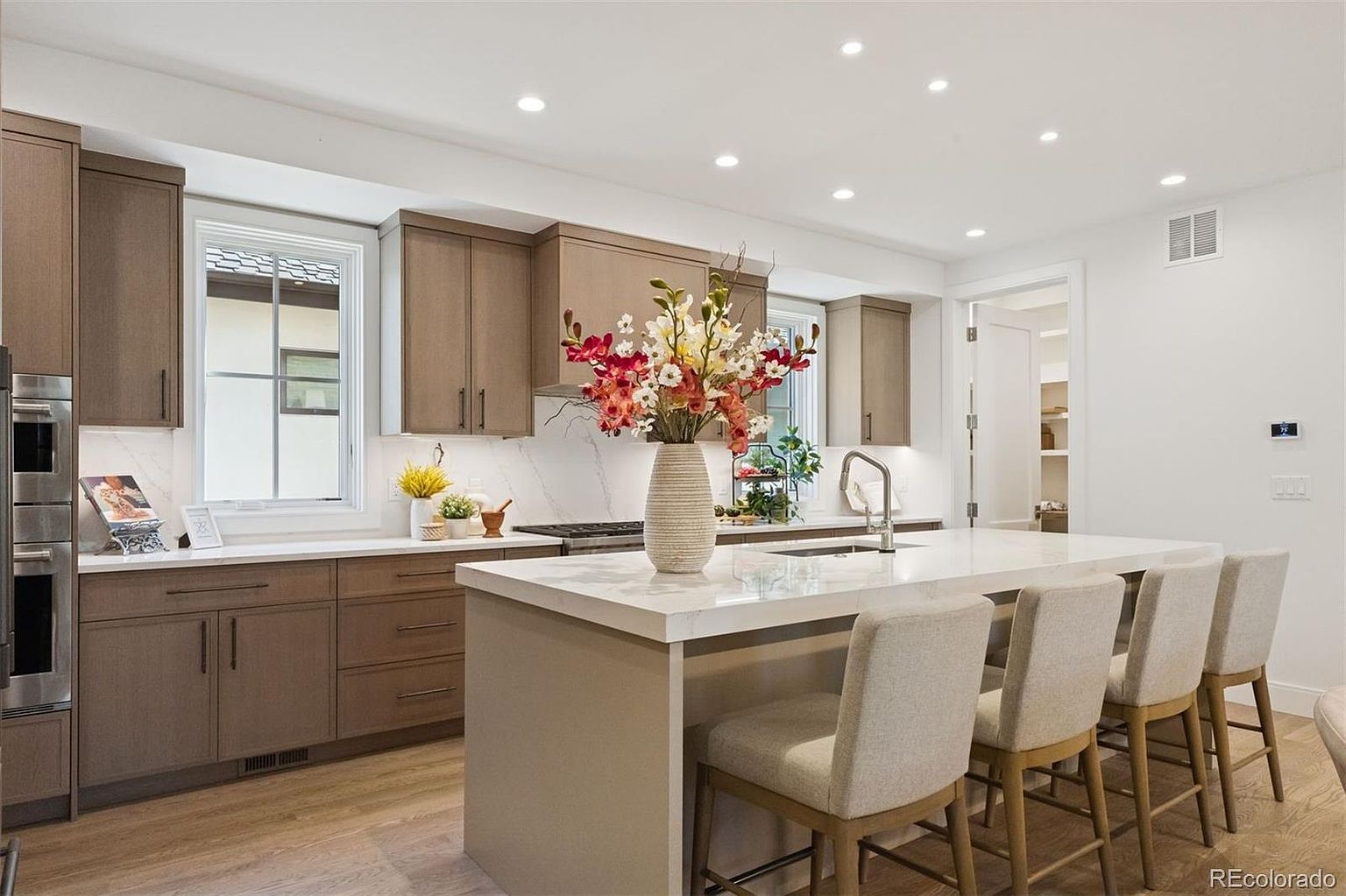 This is a well-lit kitchen featuring modern cabinetry in a neutral tone, complemented by white countertops and backsplash. A large island with seating provides ample space for food preparation and casual dining. Stainless steel appliances and recessed lighting add to the contemporary aesthetic, creating a clean and inviting space.