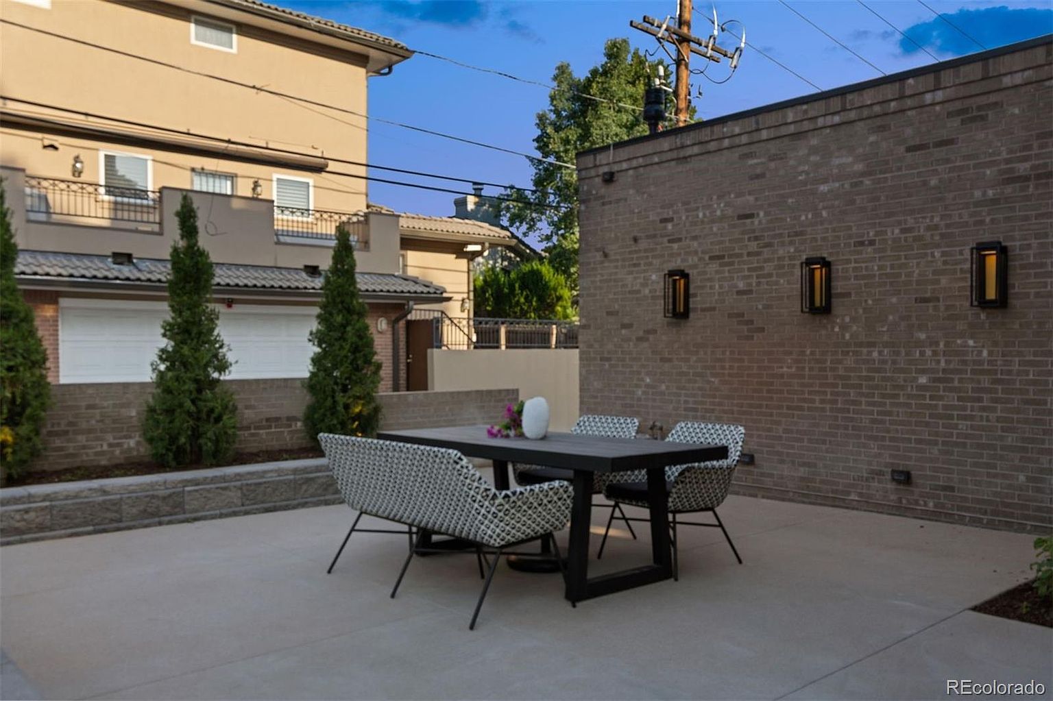 This image showcases a stylish outdoor patio area, featuring a modern dining set with a dark table and patterned chairs. The patio is paved with concrete and is enclosed by a brick wall with decorative lighting fixtures. The setting suggests a private and sophisticated outdoor living space, perfect for entertaining or relaxing.