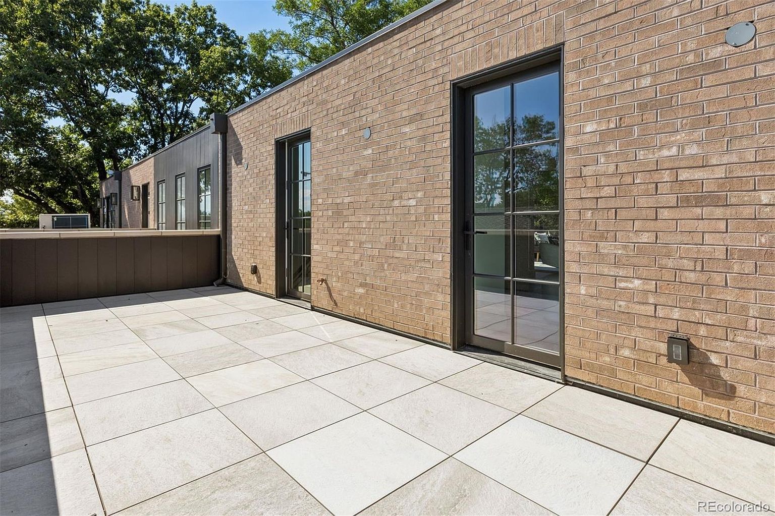 This image showcases a spacious outdoor patio or balcony area. The flooring consists of large, light-colored square tiles, creating a clean and modern aesthetic. The patio is bordered by a brick wall with large glass doors, suggesting access to the interior of the building, and a dark brown railing on the left side.