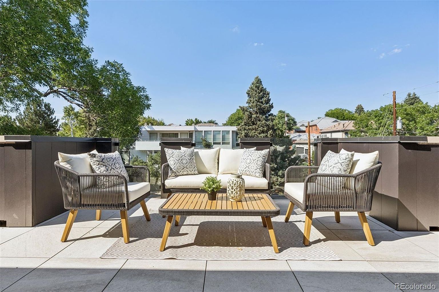 This image showcases a stylish outdoor patio or balcony setup, perfect for relaxation and entertaining. The space features a modern seating arrangement with a loveseat and two chairs, complemented by a coffee table, all set on a patterned outdoor rug. The patio is enclosed by a sleek, dark-toned railing with glass panels, offering unobstructed views of the surrounding trees and neighborhood.