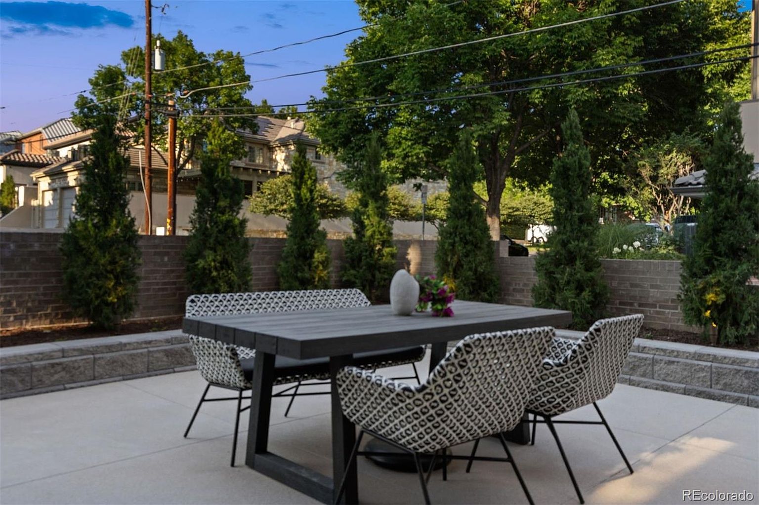 This image showcases a well-designed outdoor patio area, featuring a modern dining set with a dark-colored table and four woven chairs. The patio is paved with light-colored stone, and a low brick wall borders the space, lined with tall, slender evergreen trees. The setting suggests a private and stylish outdoor living space, perfect for entertaining or relaxing.