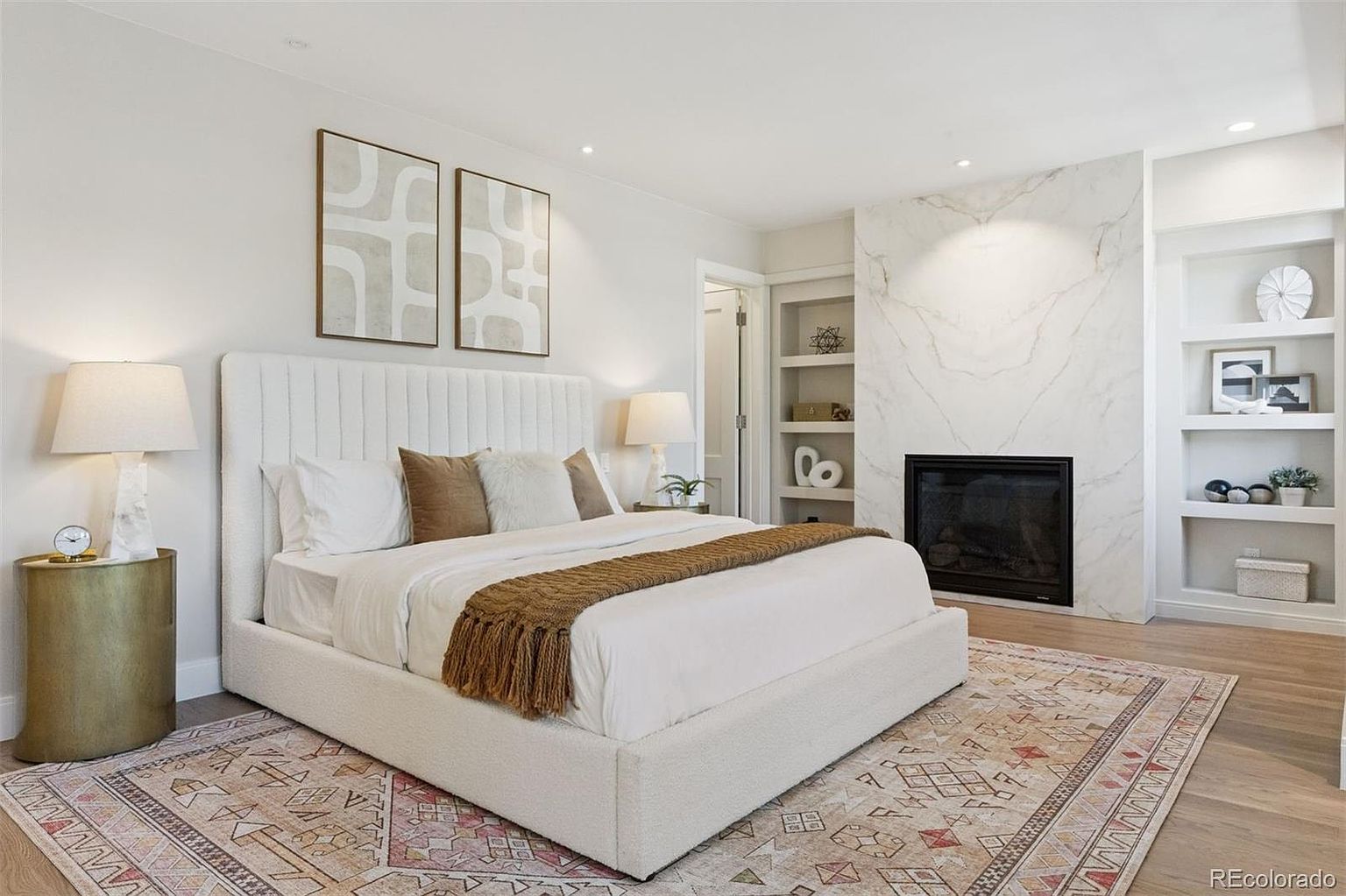 This is a primary bedroom featuring a large bed with a white upholstered headboard and base, complemented by neutral-toned pillows and a brown throw. The room includes a fireplace with a marble surround and built-in shelving on either side, adding architectural interest. The space is decorated with modern artwork and a patterned area rug, creating a serene and stylish atmosphere.