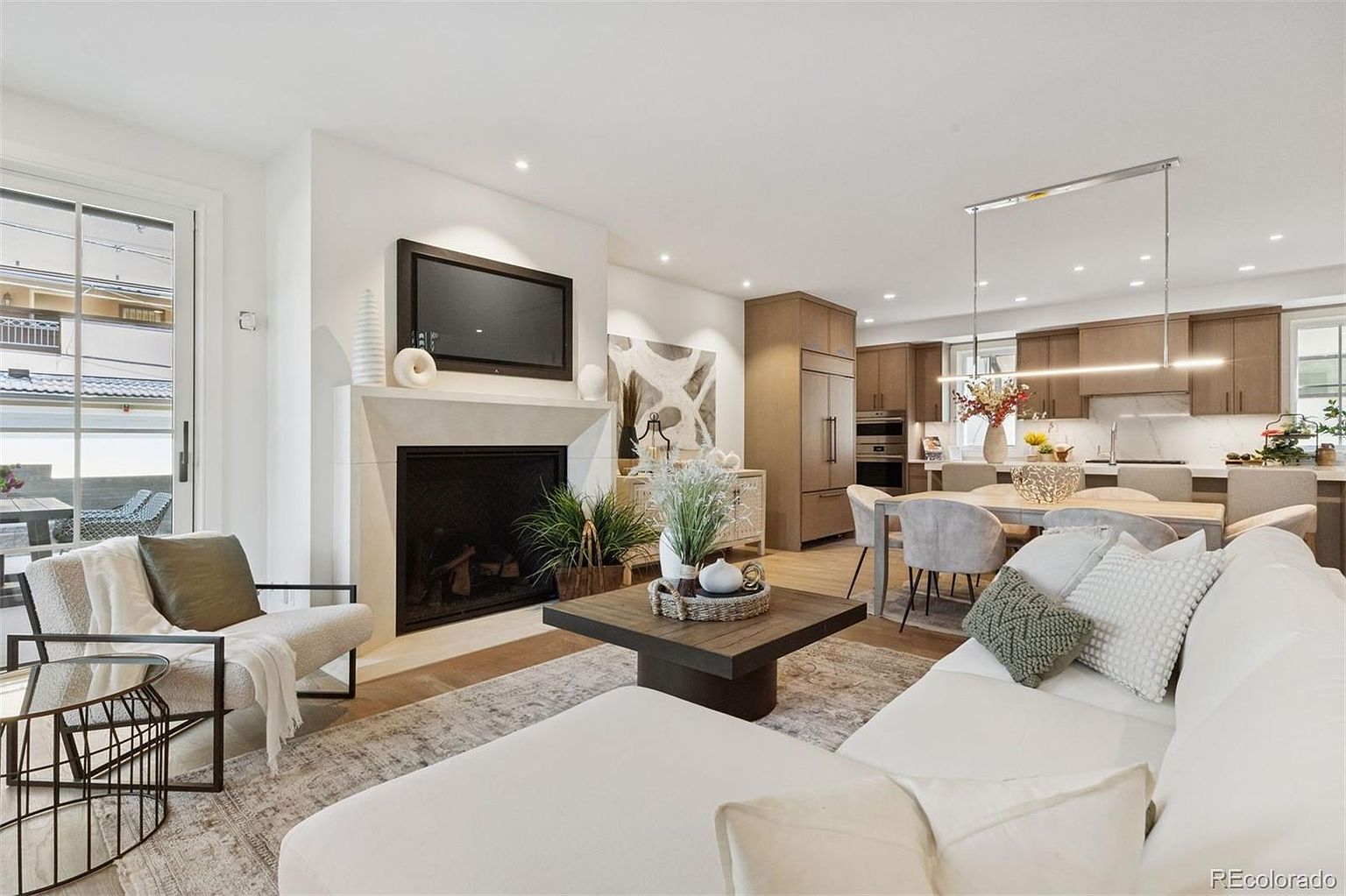 This is an interior shot of a modern living room that seamlessly transitions into a kitchen and dining area. The living room features a fireplace with a mounted television, a comfortable armchair, and a sectional sofa. The open-concept design creates a spacious and inviting atmosphere, perfect for entertaining and family living.