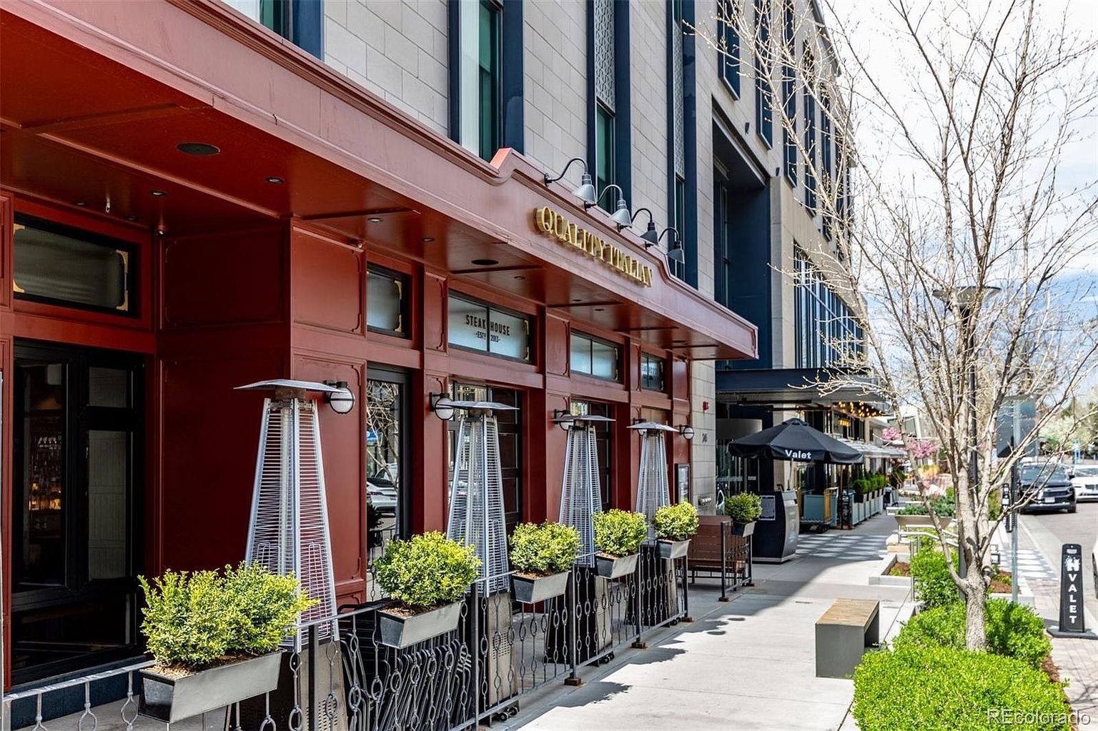 This is a street-level view of a building featuring a restaurant called "Quality Italian Steak House". The restaurant has a dark red facade with gold lettering and is lined with outdoor heaters and planters. The sidewalk is clean and well-maintained, with a valet stand visible in the background, suggesting a high-end establishment.