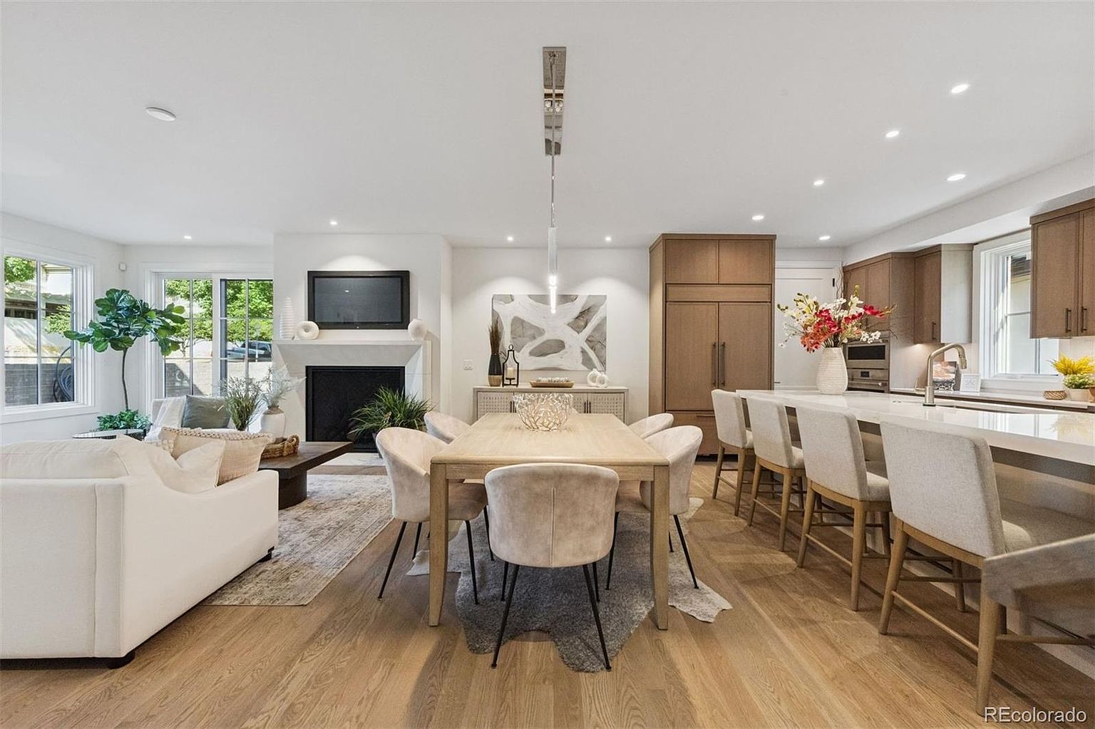 This is an interior shot showcasing an open-concept living space that seamlessly integrates the living room, dining area, and kitchen. The living room features a comfortable white sofa, a fireplace with a mounted TV, and large windows providing natural light. The dining area includes a wooden table with upholstered chairs and a modern chandelier, while the kitchen boasts a large island with bar seating and modern cabinetry. The overall impression is one of a bright, airy, and stylish home perfect for modern living.