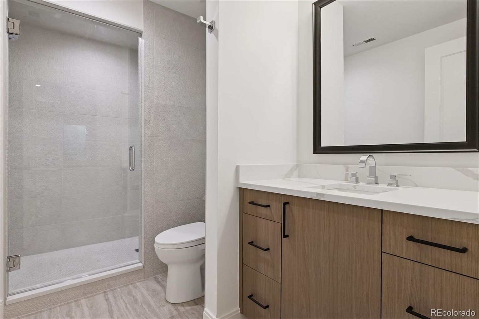 This is a well-lit bathroom featuring a glass-enclosed shower, a modern toilet, and a vanity with a white countertop and wood-grain cabinets. A black-framed mirror hangs above the sink, and the flooring appears to be a light-colored tile. The overall impression is clean, contemporary, and functional.