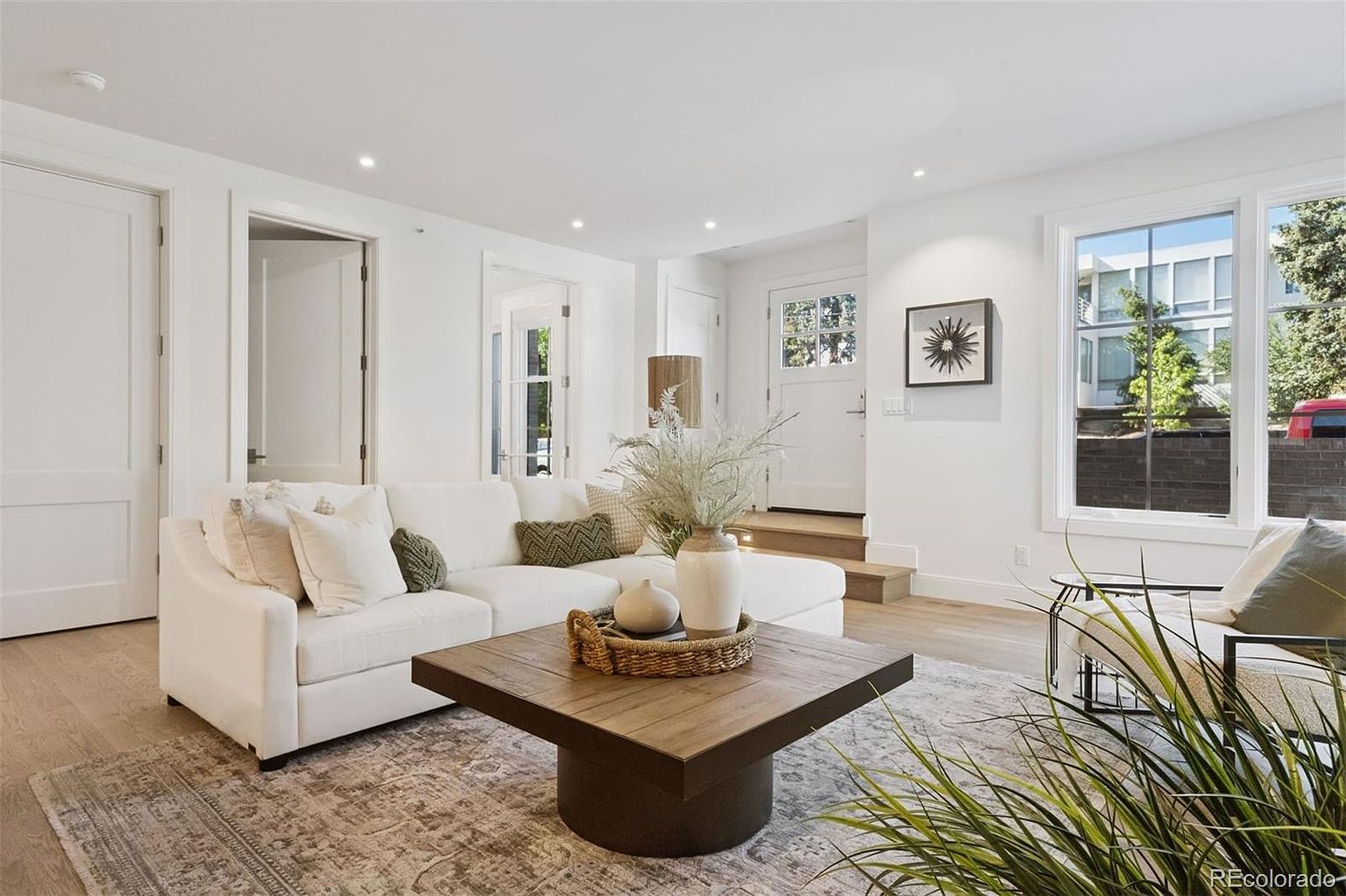 This is a bright and airy living room featuring a white sectional sofa with decorative pillows, a square wooden coffee table with a woven tray and vases, and a neutral-toned area rug. The room has white walls, recessed lighting, and a large window that provides natural light. The overall style is modern and inviting.