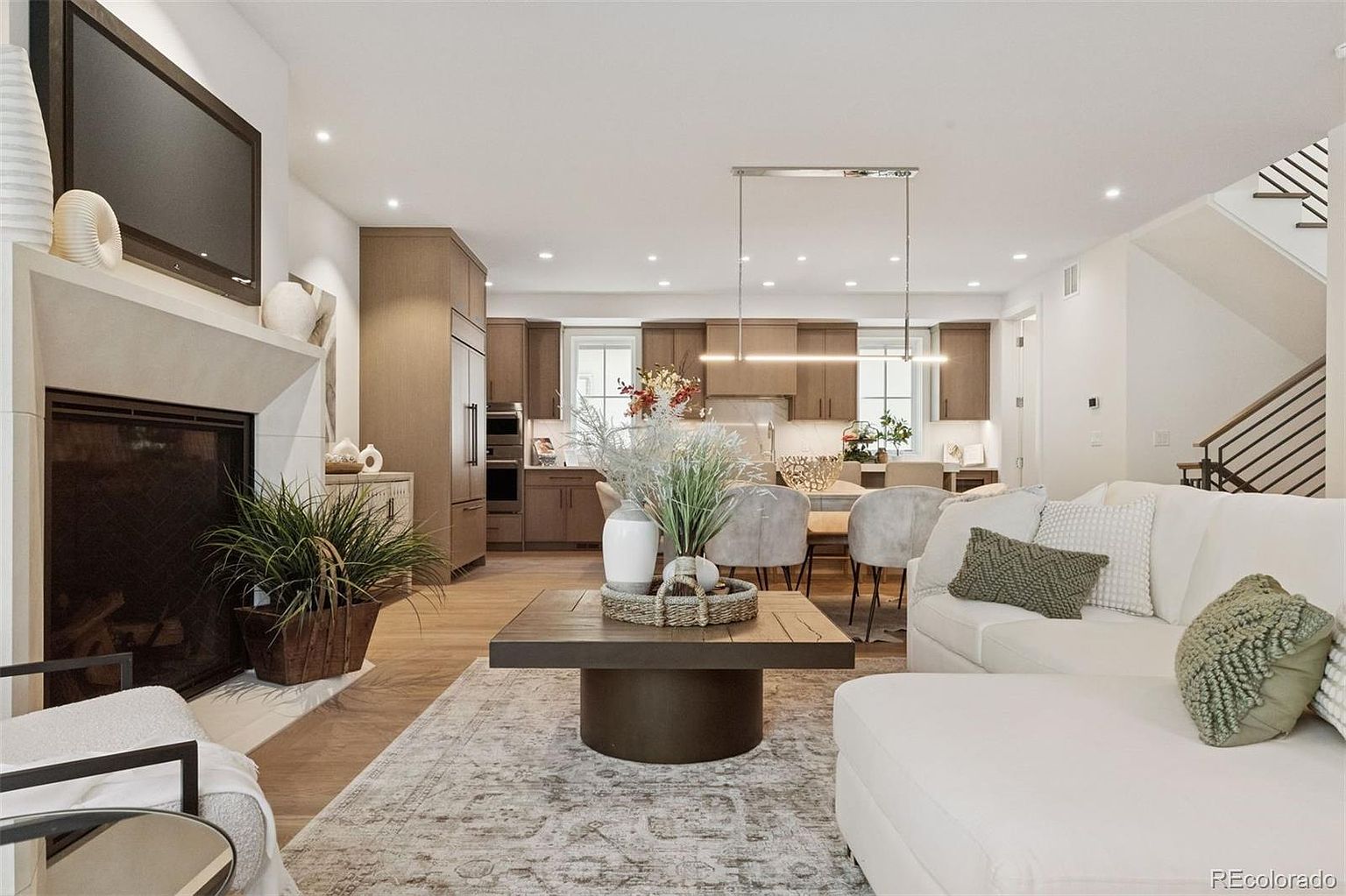 This is an interior shot of a modern living room that seamlessly flows into the kitchen and dining area. The living room features a fireplace with a mounted TV, a comfortable white sectional sofa with decorative pillows, and a stylish coffee table on a patterned rug. The open-concept design enhances the sense of space and connectivity, making it ideal for entertaining and family living.