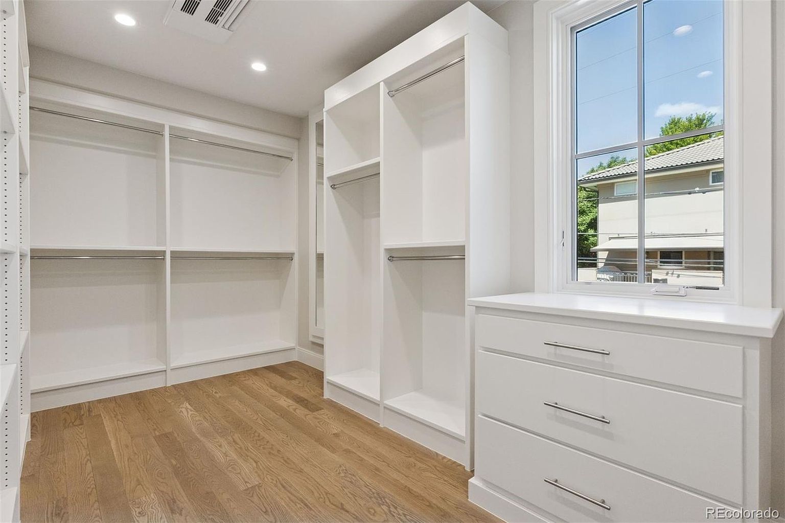 This is a well-organized walk-in closet featuring custom white shelving and drawers. The closet includes multiple hanging rods and adjustable shelves for versatile storage options. A window provides natural light, enhancing the bright and clean aesthetic of the space, and the hardwood flooring adds a touch of warmth.