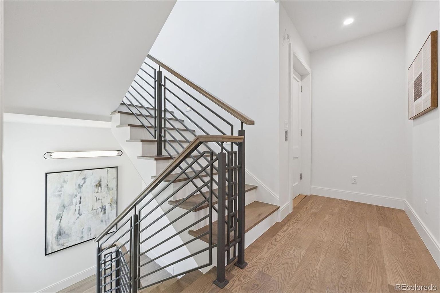 This interior shot showcases a modern hallway and staircase. The staircase features wooden steps, white risers, and a metal railing with wooden accents. The hallway has light wood flooring and white walls, creating a bright and clean aesthetic, complemented by artwork and a minimalist light fixture.