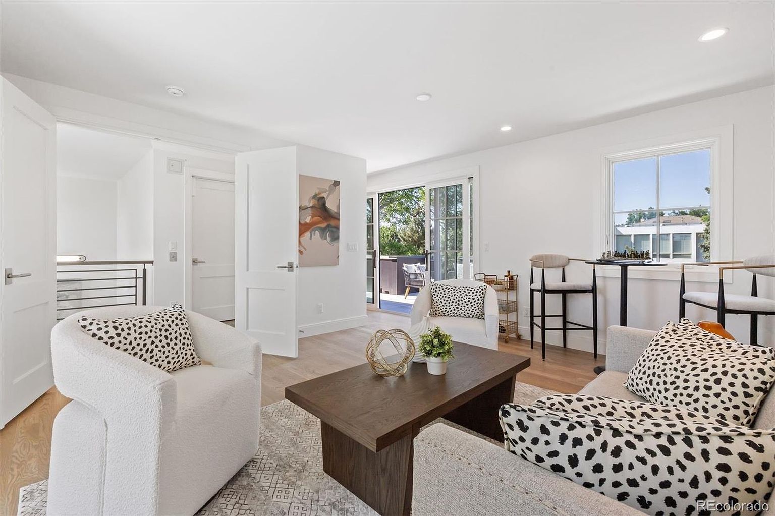 This is an interior shot of a living room featuring a light wood floor, white walls, and neutral-toned furniture. The room is well-lit with natural light coming through a window and sliding glass doors that lead to a balcony. The furniture includes a sofa and two armchairs with patterned pillows, and a dark wood coffee table sits in the center of the room.