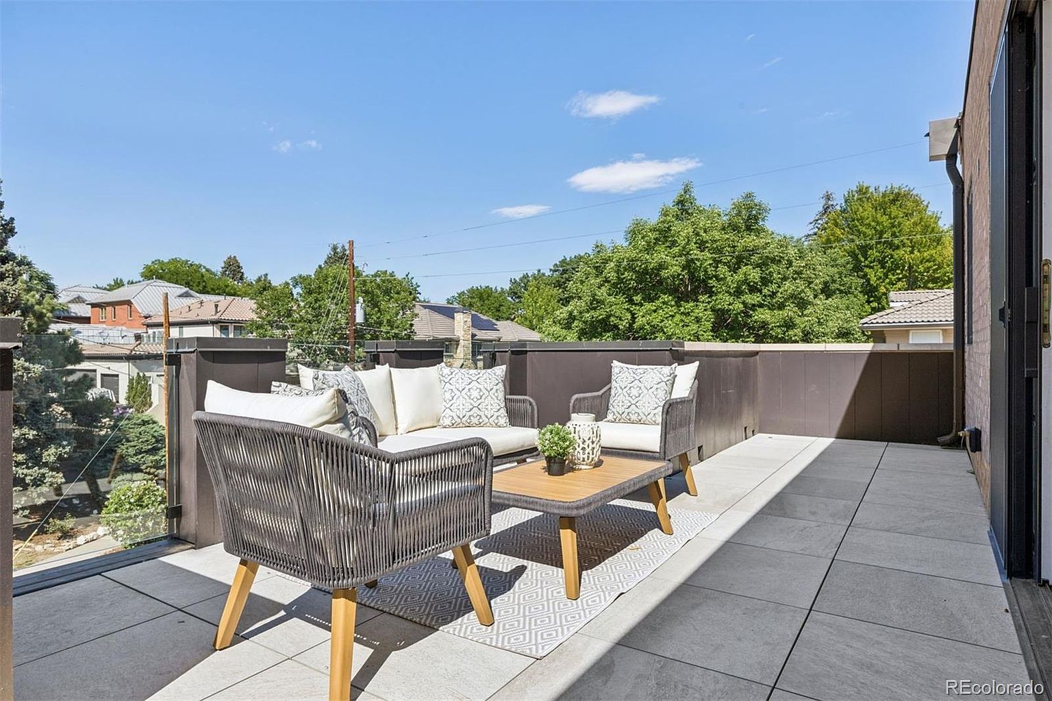 This image showcases a stylish outdoor patio or balcony area, furnished with a modern seating arrangement including a sofa and chairs with woven detailing, complemented by a wooden coffee table. The space is adorned with neutral-toned cushions and a patterned rug, creating a cozy and inviting atmosphere. The patio features tiled flooring and a glass railing, offering a clear view of the surrounding neighborhood and sky.