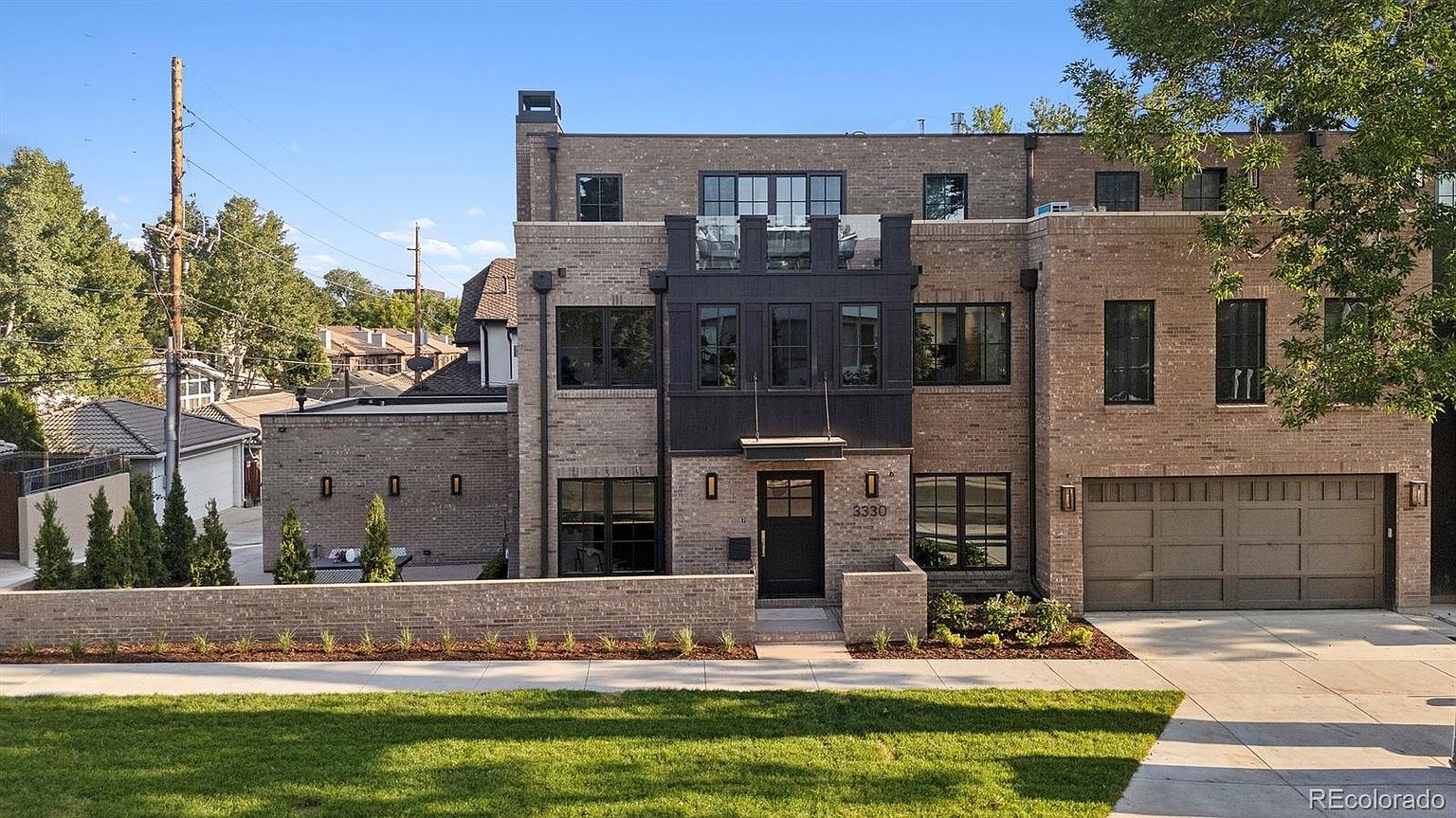 The image showcases the front exterior of a modern brick home. The house features a combination of light brown brick and dark metal accents around the windows and entryway. A well-manicured lawn and a low brick wall enhance the property's curb appeal, while a paved driveway leads to an attached garage on the right side of the house.