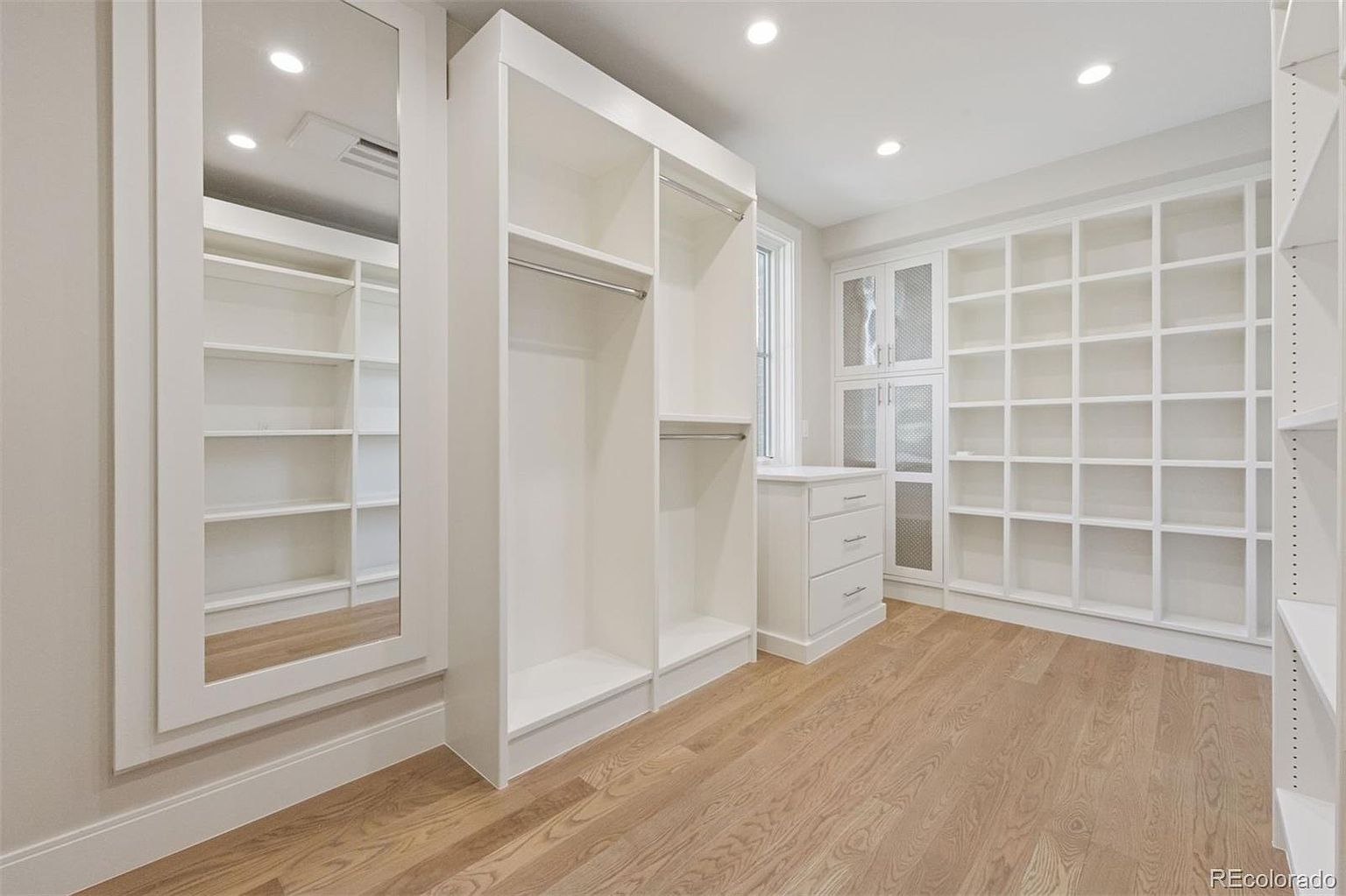 This is a well-organized walk-in closet featuring custom white shelving and hanging rods. The closet is spacious with light wood flooring and ample storage options, including drawers and cubbies. The design is clean and modern, maximizing space and functionality.