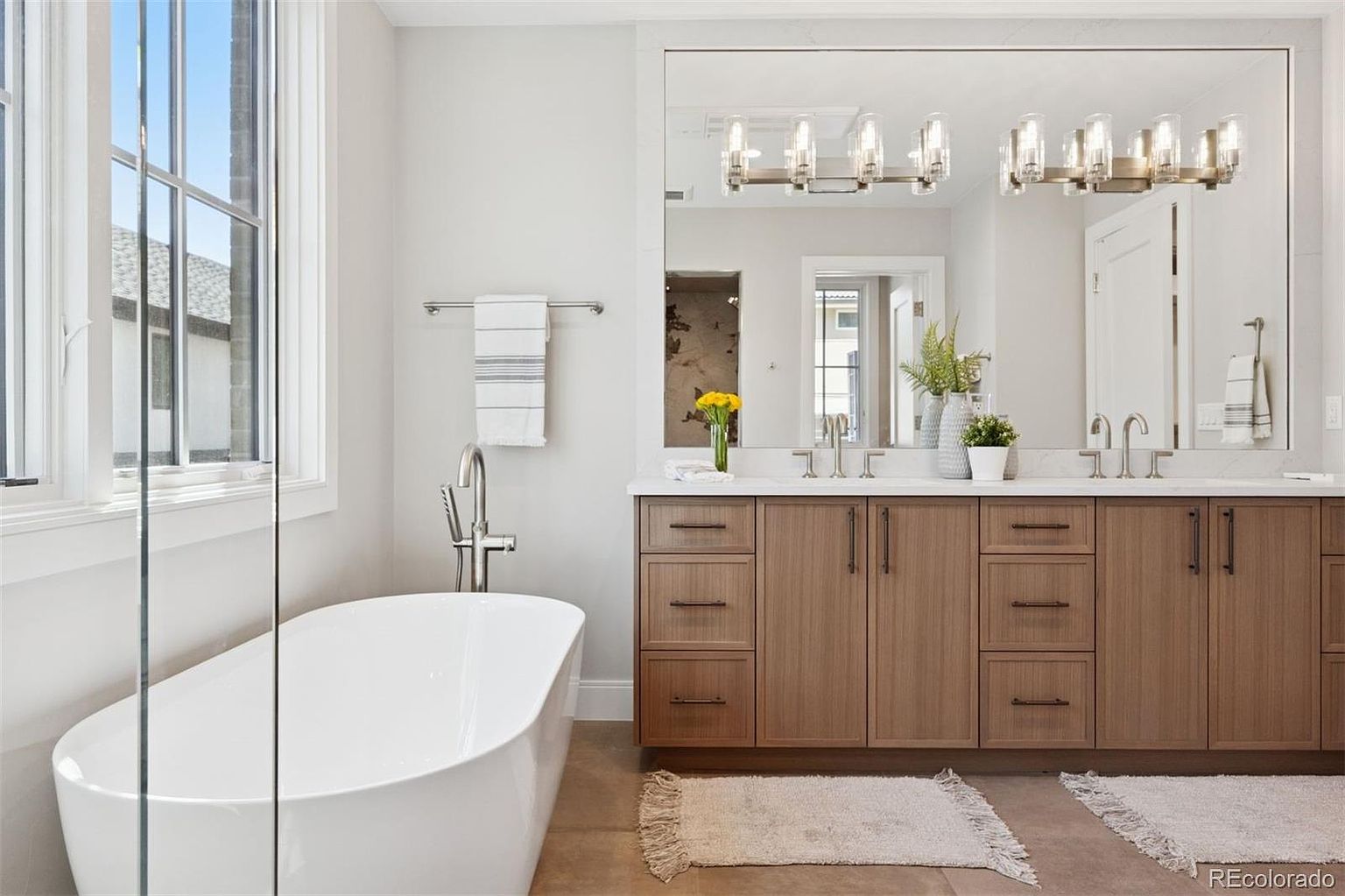 This is a bright and modern primary bathroom featuring a freestanding white bathtub next to a large window, a double vanity with wood-tone cabinets, and a large mirror with stylish lighting. The space is clean and well-lit, creating a spa-like atmosphere, and the neutral color palette enhances the sense of tranquility and luxury.