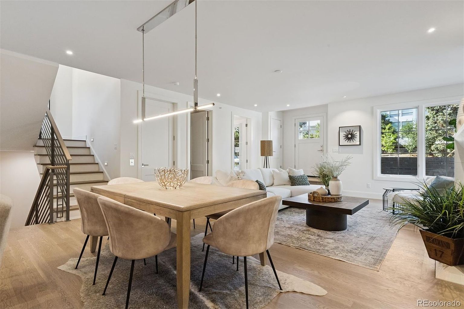 This is an interior shot showcasing a combined dining and living area. The dining area features a wooden table with modern chairs, illuminated by a sleek, linear pendant light. The living area includes a comfortable white sofa, a dark coffee table, and a large window providing natural light, creating a bright and inviting atmosphere.