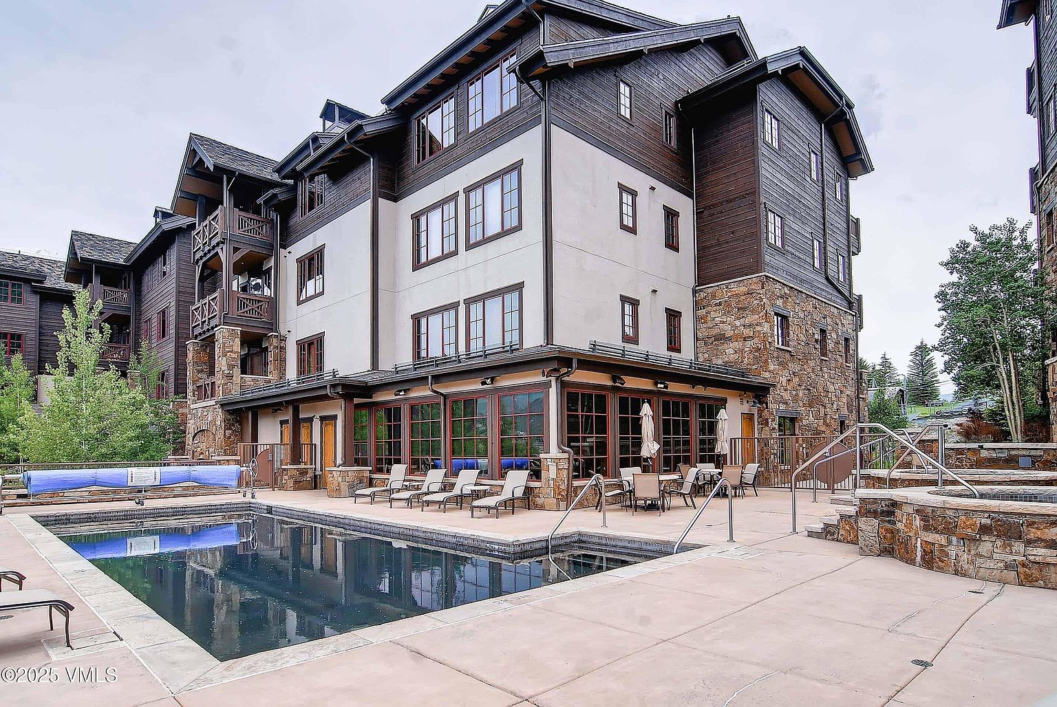 This image showcases a luxurious outdoor pool and spa area of a multi-story building. The pool's dark water reflects the building's architecture, which features a blend of stone, wood, and stucco. Lounge chairs and tables are arranged around the pool, suggesting a relaxing and upscale environment.