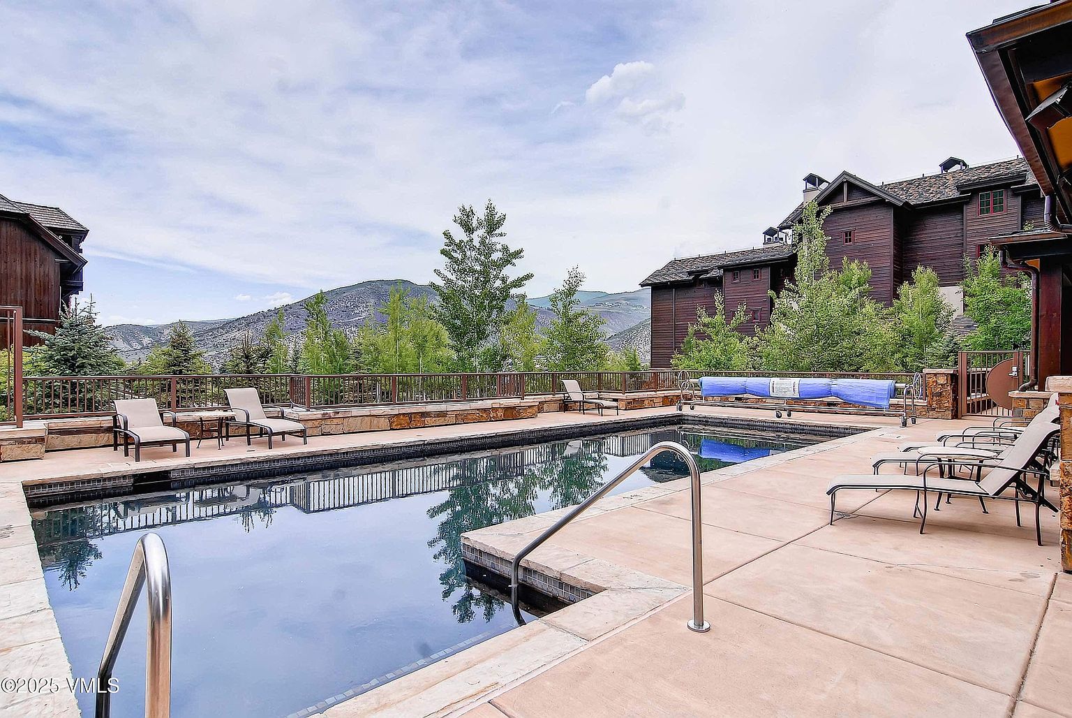 This image showcases a luxurious outdoor pool area with lounge chairs and a view of the mountains. The pool is surrounded by a stone patio and a wooden fence, with a large wooden house in the background. The scene evokes a sense of relaxation and upscale living.