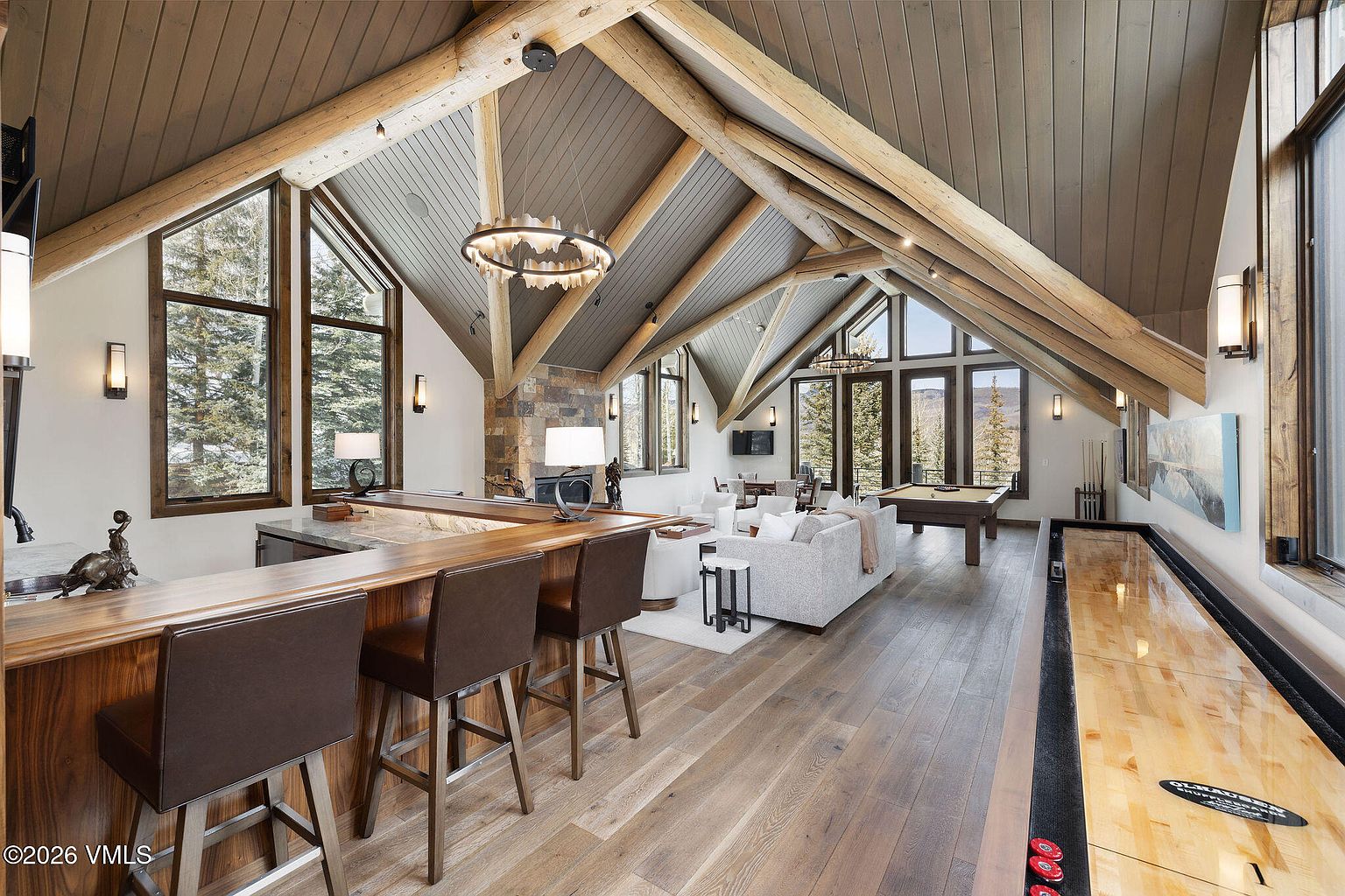 This is a spacious living room featuring a high vaulted ceiling with exposed wooden beams, creating a rustic yet elegant atmosphere. The room includes a bar area with seating, a stone fireplace, a comfortable seating area with a sofa and chairs, a pool table, and a shuffleboard table, making it an ideal space for relaxation and entertainment. Large windows provide ample natural light and views of the surrounding landscape.