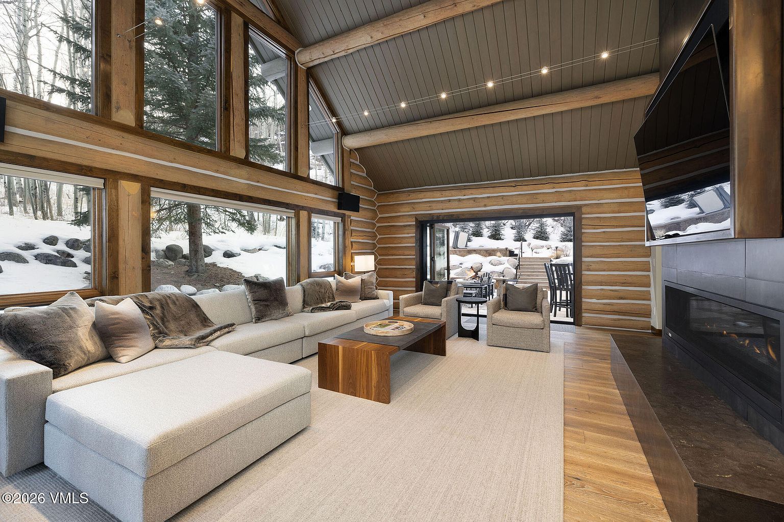 This is a spacious living room with a rustic yet luxurious feel, featuring log walls, a high vaulted ceiling with exposed beams, and large windows offering views of a snowy landscape. A large sectional sofa anchors the room, complemented by armchairs, a wooden coffee table, and a fireplace with a mounted television. The room is well-lit and inviting, creating a cozy atmosphere.