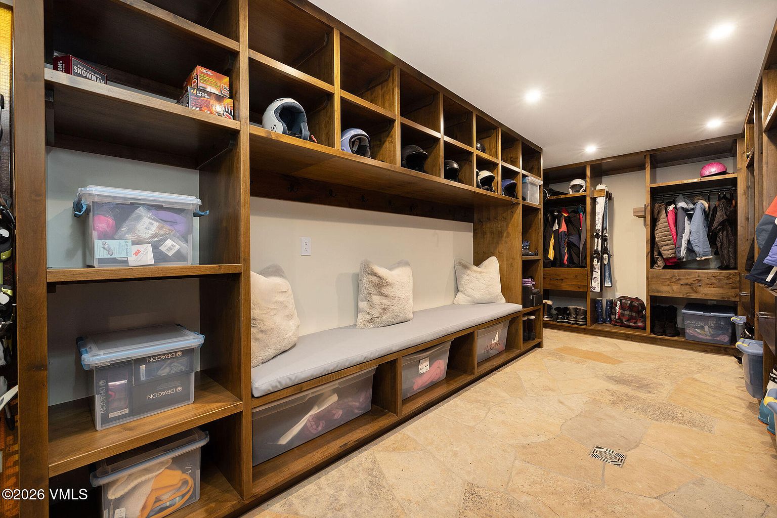 This is a well-organized walk-in closet featuring custom wooden shelving and storage solutions. The closet includes open shelving for helmets and bins, a bench with storage underneath, and hanging rods for clothing. The flooring is a light-colored stone tile, and the overall impression is one of functionality and ample storage space.