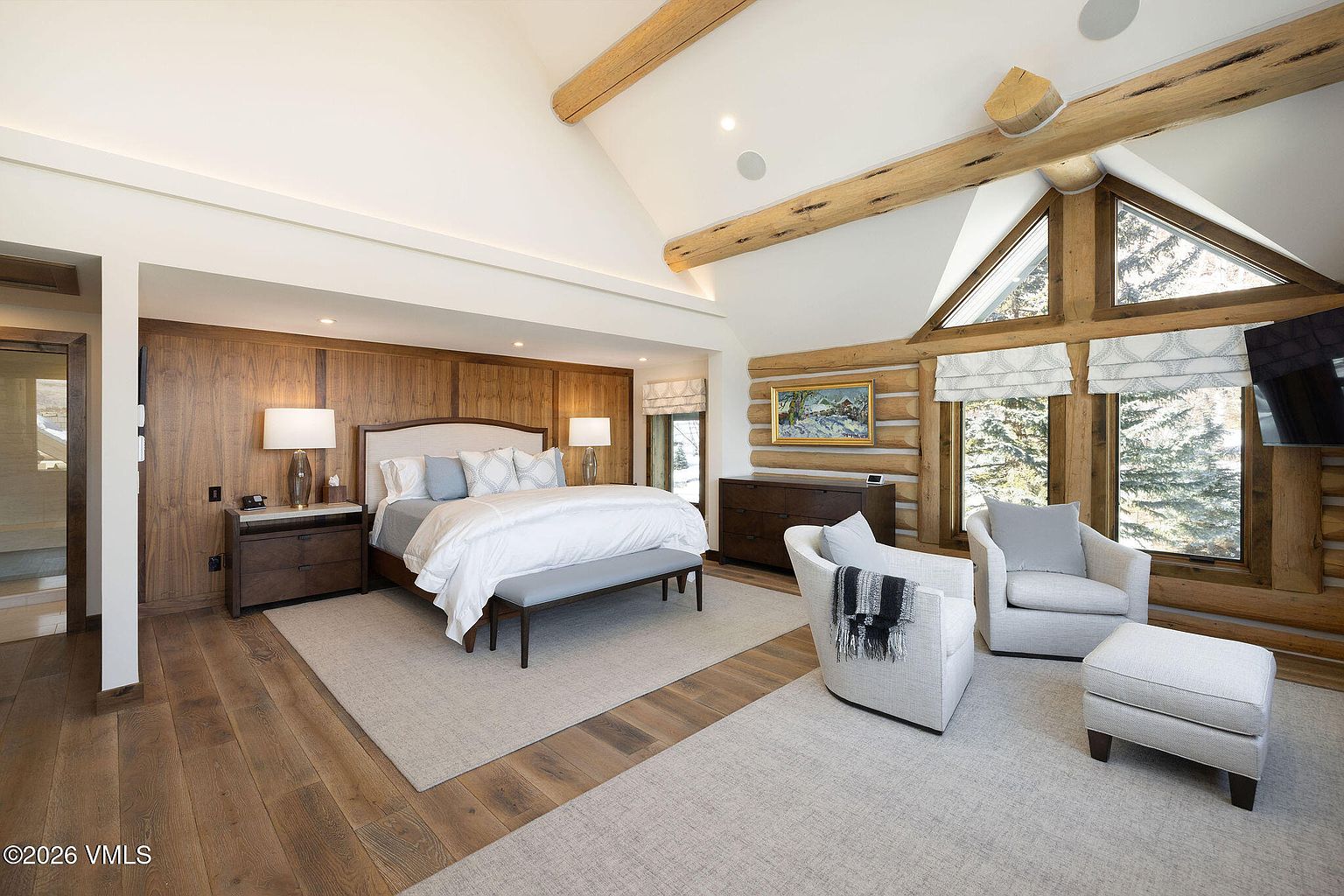 This is a primary bedroom featuring a king-size bed with a white duvet and light blue pillows, flanked by wooden nightstands and lamps. The room has wood-paneled walls behind the bed and a vaulted ceiling with exposed wooden beams, creating a rustic yet luxurious ambiance. A seating area with two armchairs and an ottoman is positioned near a window, offering a cozy retreat within the bedroom.