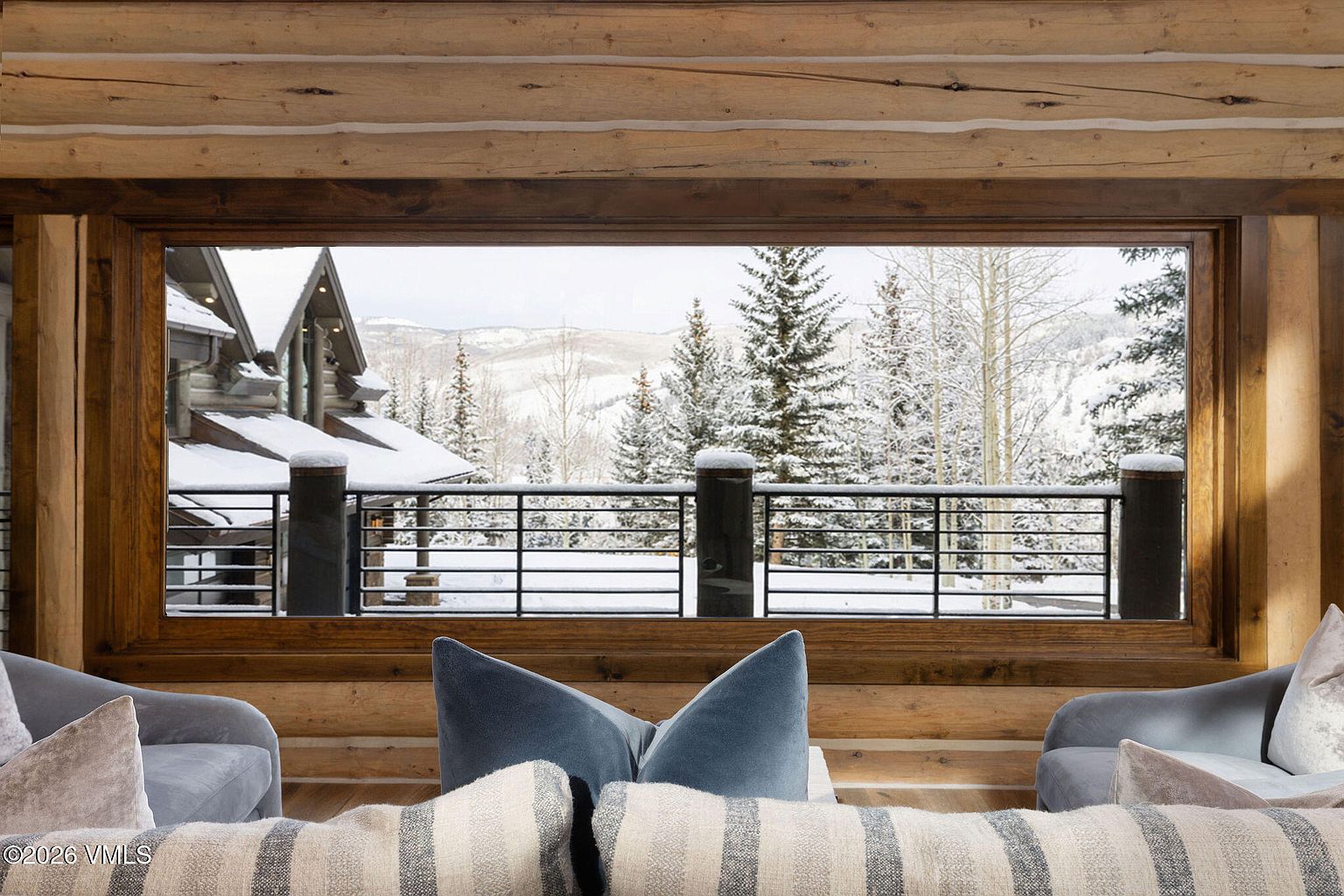 This interior shot showcases a cozy living room with a large window framing a snowy mountain landscape. The room features rustic wooden walls and beams, complemented by comfortable seating with plush pillows. The view outside adds a sense of serenity and connection to nature, enhancing the room's appeal.