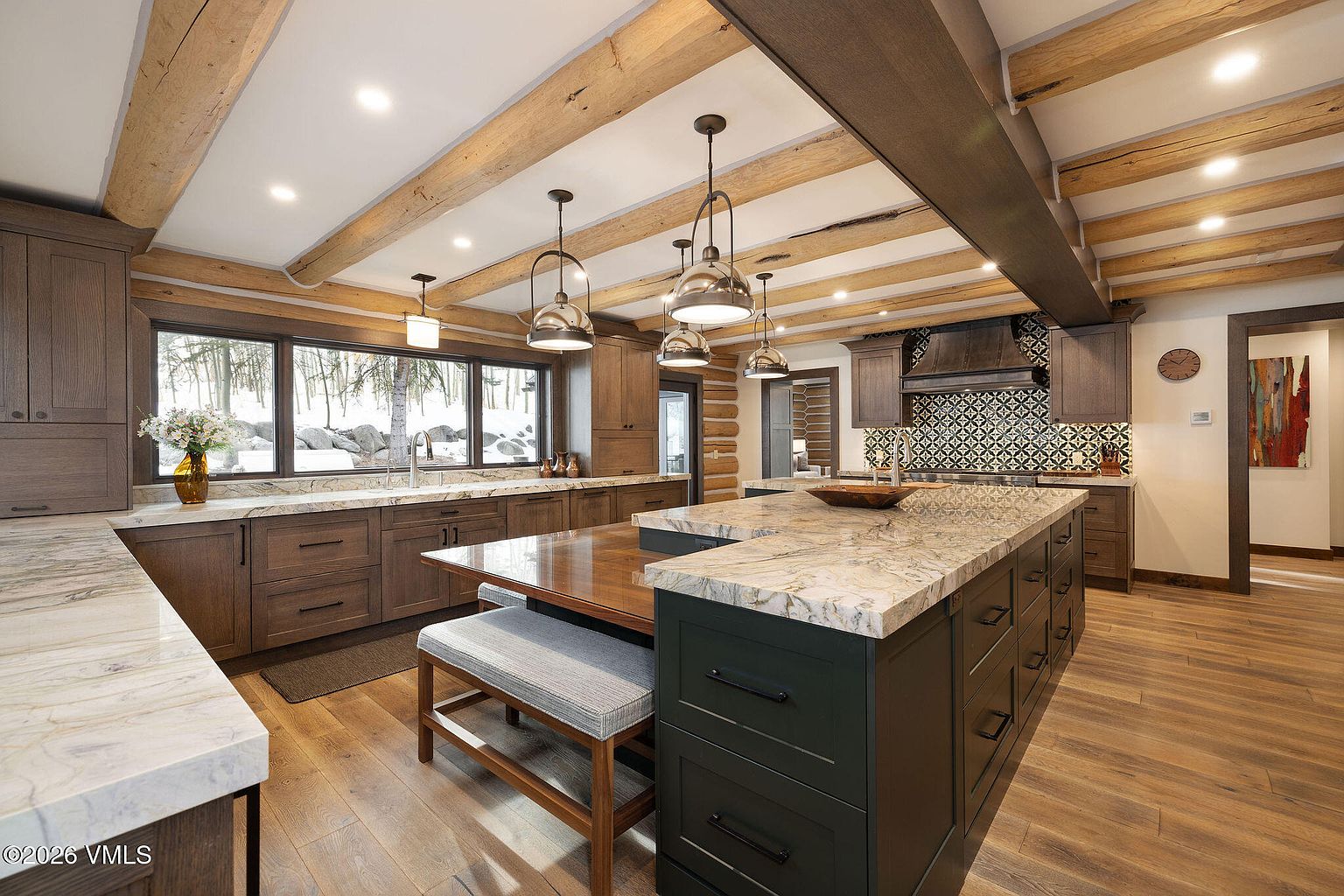 This is a well-lit kitchen with a rustic yet modern design. It features wooden beams on the ceiling, dark wood cabinetry, and a large island with a granite countertop and a built-in wooden table with a bench. The kitchen also has a window overlooking a snowy landscape, adding to the cozy atmosphere.