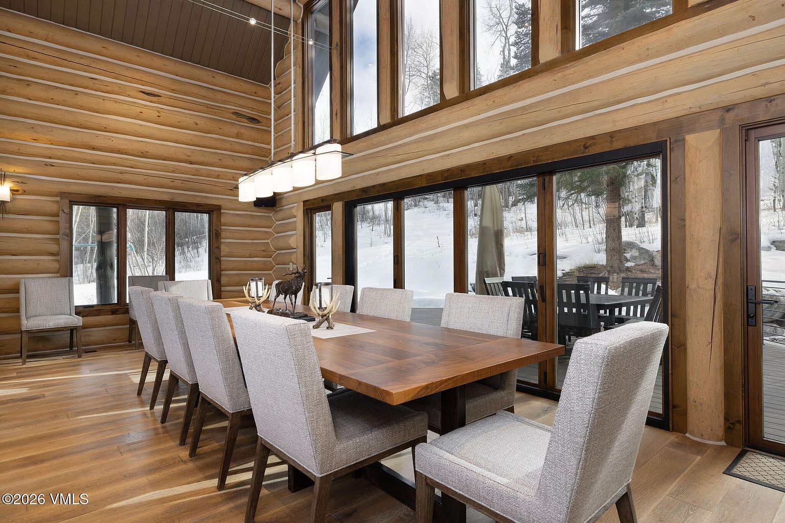 This is an interior shot of a dining room featuring a large wooden table surrounded by upholstered chairs. The room is characterized by its log cabin style, with wooden walls and large windows that offer a view of a snowy landscape. A modern chandelier hangs above the table, blending rustic and contemporary elements.