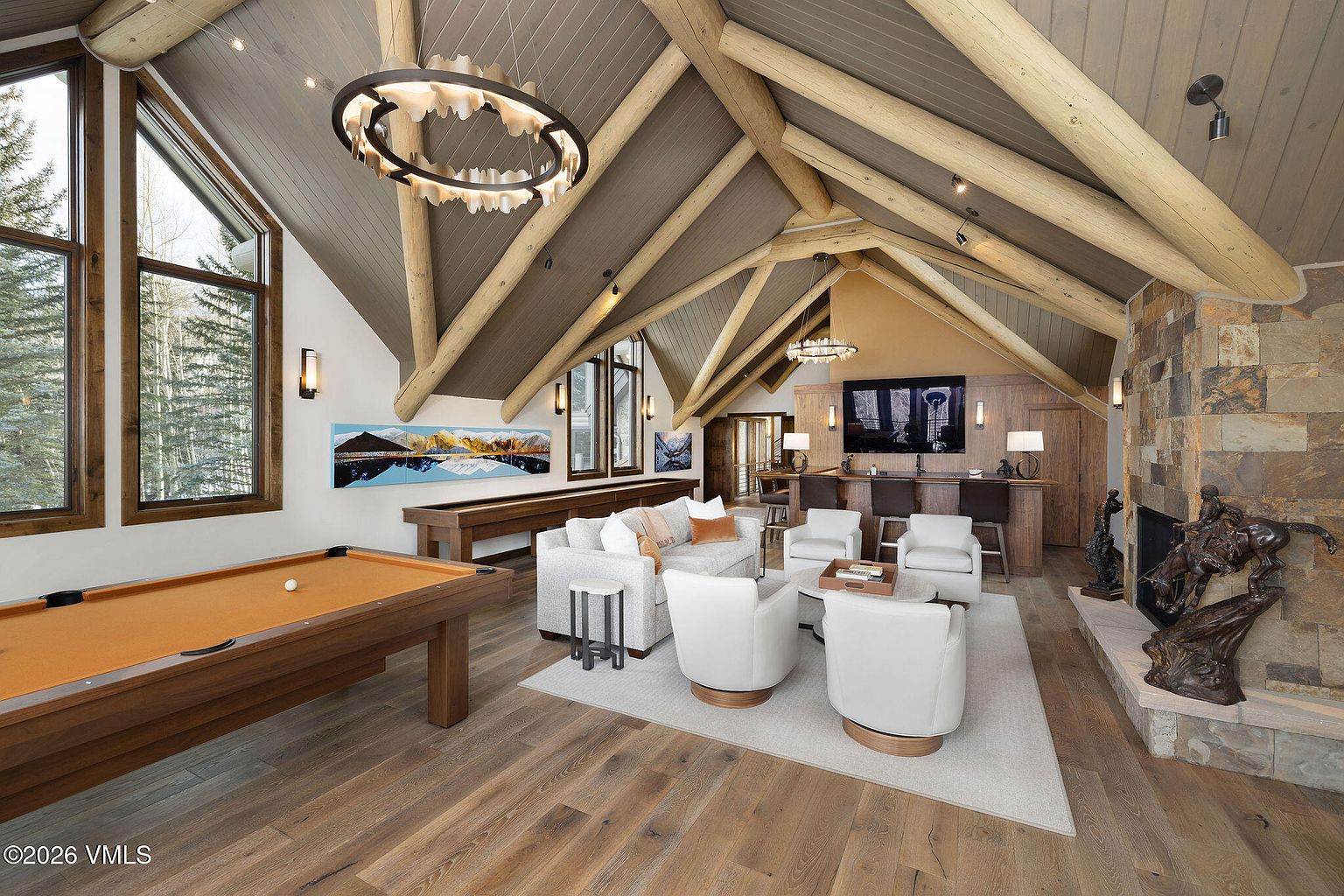 This is an interior shot of a luxurious living room featuring a high vaulted ceiling with exposed wooden beams. The room includes a pool table, a shuffleboard table, a seating area with white swivel chairs and a sofa, and a stone fireplace with bronze sculptures. The overall impression is one of rustic elegance and spaciousness.