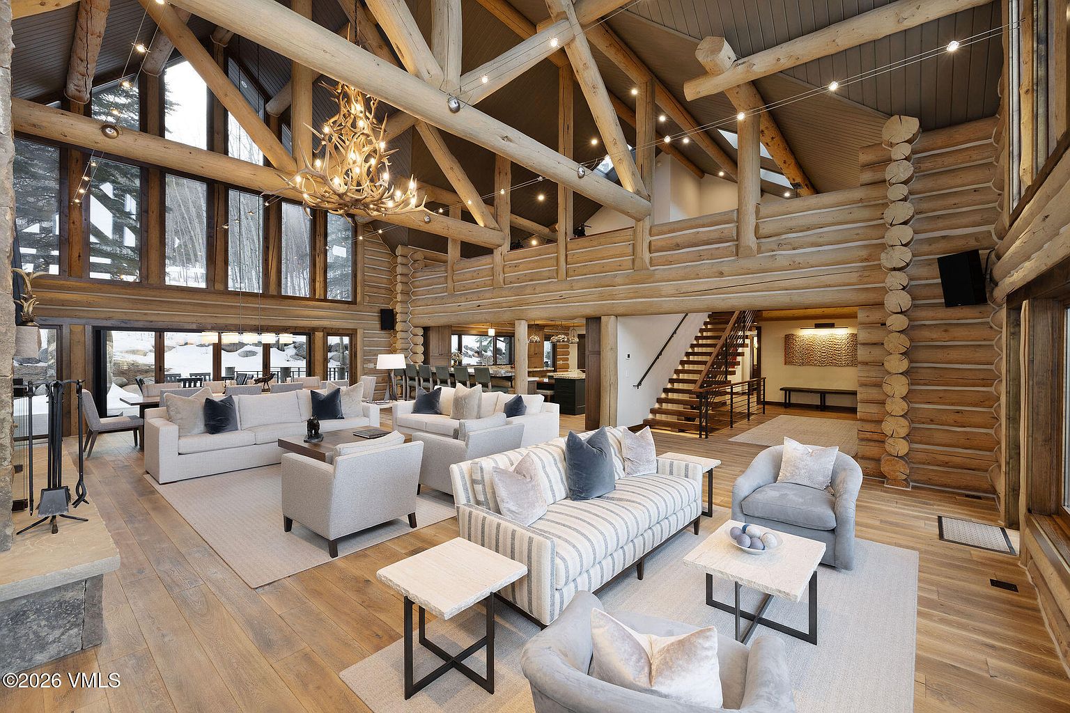 This is a spacious living room in a log cabin style home, featuring exposed wooden beams, a large antler chandelier, and multiple seating areas with plush sofas and armchairs. The room is well-lit with natural light from large windows, and the open floor plan connects to other areas of the house, creating a warm and inviting atmosphere.