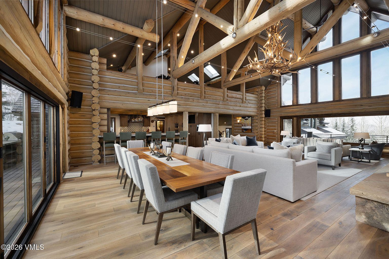 This is a spacious, luxurious living and dining area featuring a log cabin design with exposed wooden beams and a high ceiling. A large wooden dining table with gray upholstered chairs sits adjacent to a comfortable living area with a sectional sofa and armchairs. Natural light floods the space through large windows, showcasing a scenic outdoor view, and an antler chandelier adds a rustic touch.