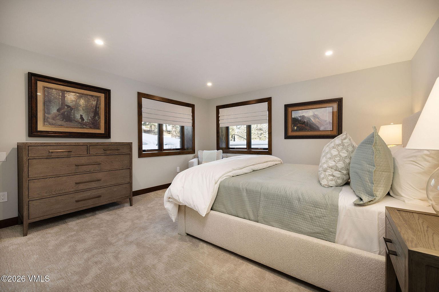 This is a well-lit guest bedroom featuring neutral tones and comfortable furnishings. The room includes a bed with a light green quilt and decorative pillows, a wooden dresser with artwork above, and two windows with roman shades. The overall impression is cozy and inviting, perfect for a relaxing stay.