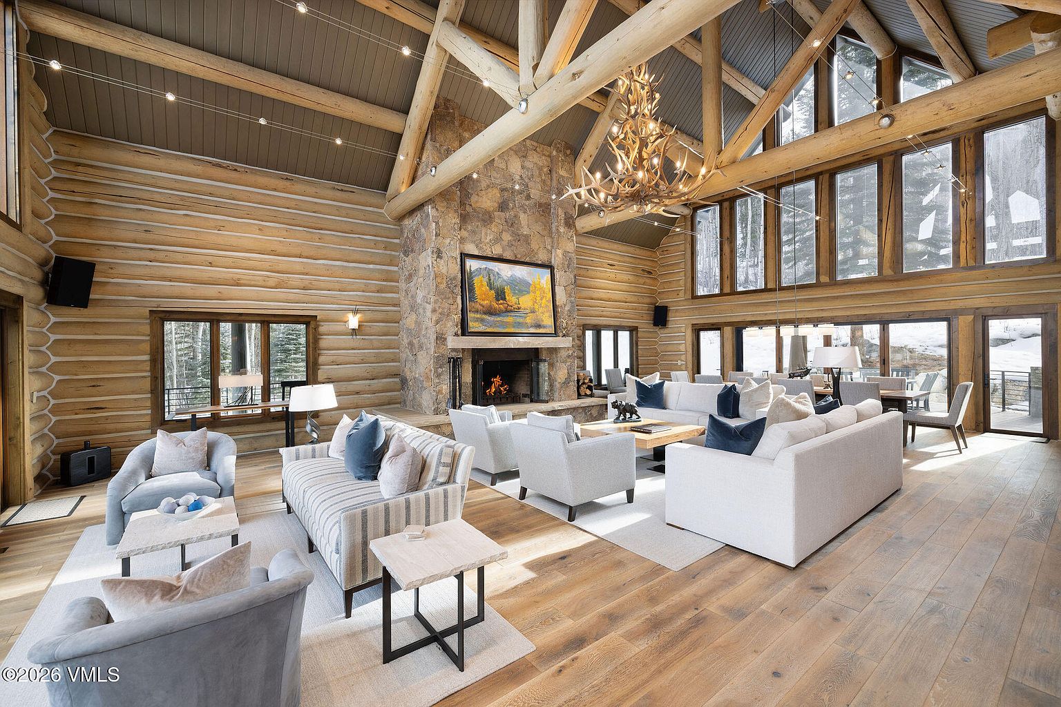 This is a spacious living room featuring a high vaulted ceiling with exposed wooden beams and log walls, creating a rustic yet luxurious atmosphere. A large stone fireplace serves as a focal point, complemented by comfortable seating arrangements including sofas and armchairs. Large windows provide ample natural light and views of the surrounding landscape, enhancing the room's appeal for relaxation and entertainment.