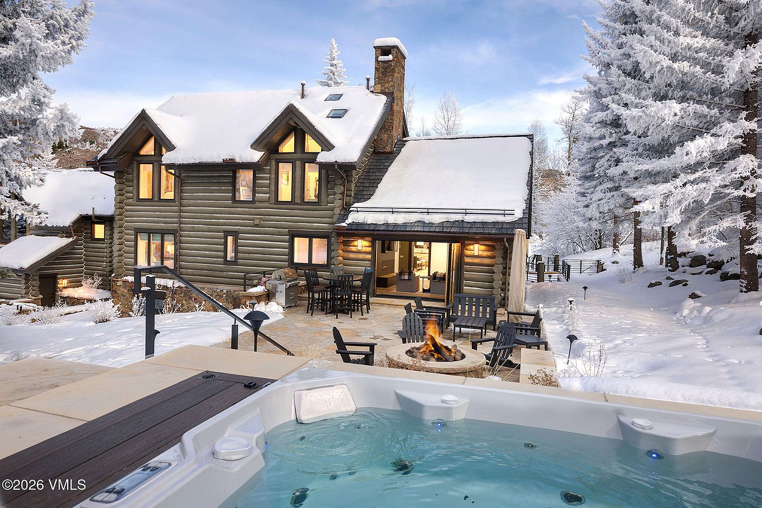This image showcases the rear exterior of a log cabin home in a snowy, winter setting. A hot tub with clear blue water is in the foreground, with a stone patio featuring a fire pit and outdoor seating. The log cabin has snow-covered roofs and illuminated windows, creating a warm and inviting atmosphere.