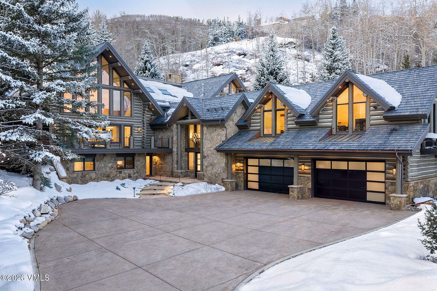 This is a front exterior view of a luxurious mountain home. The house features a combination of stone and wood siding, with large windows that offer stunning views. A spacious driveway leads up to a two-car garage, and the surrounding landscape is covered in snow, creating a serene and picturesque setting.