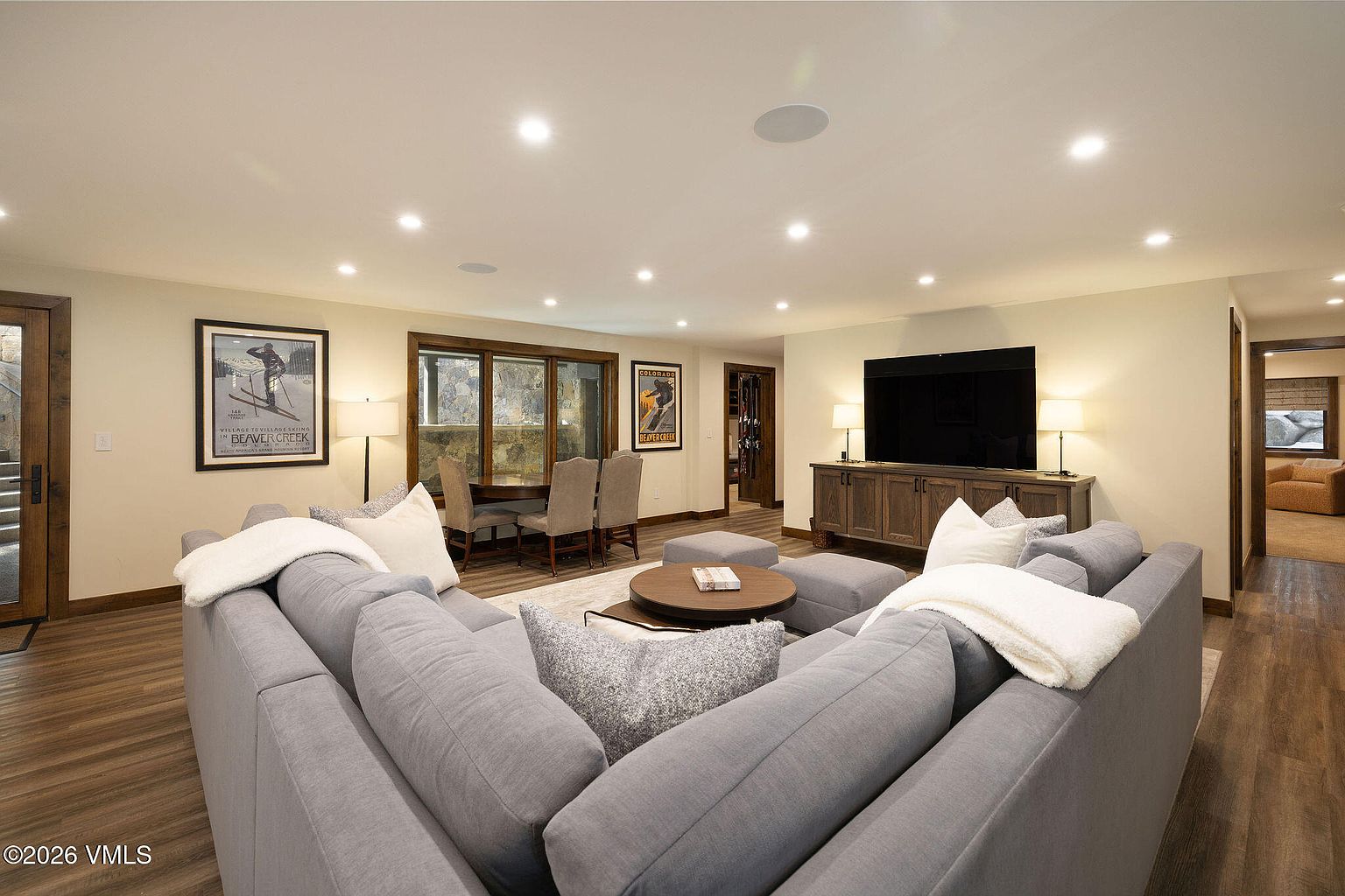 This is an interior shot of a living room featuring a large, gray sectional sofa with throw pillows and blankets. A round coffee table sits in the center of the room, and a media console with a large television is against the wall. The room has recessed lighting and wood flooring, creating a warm and inviting atmosphere.