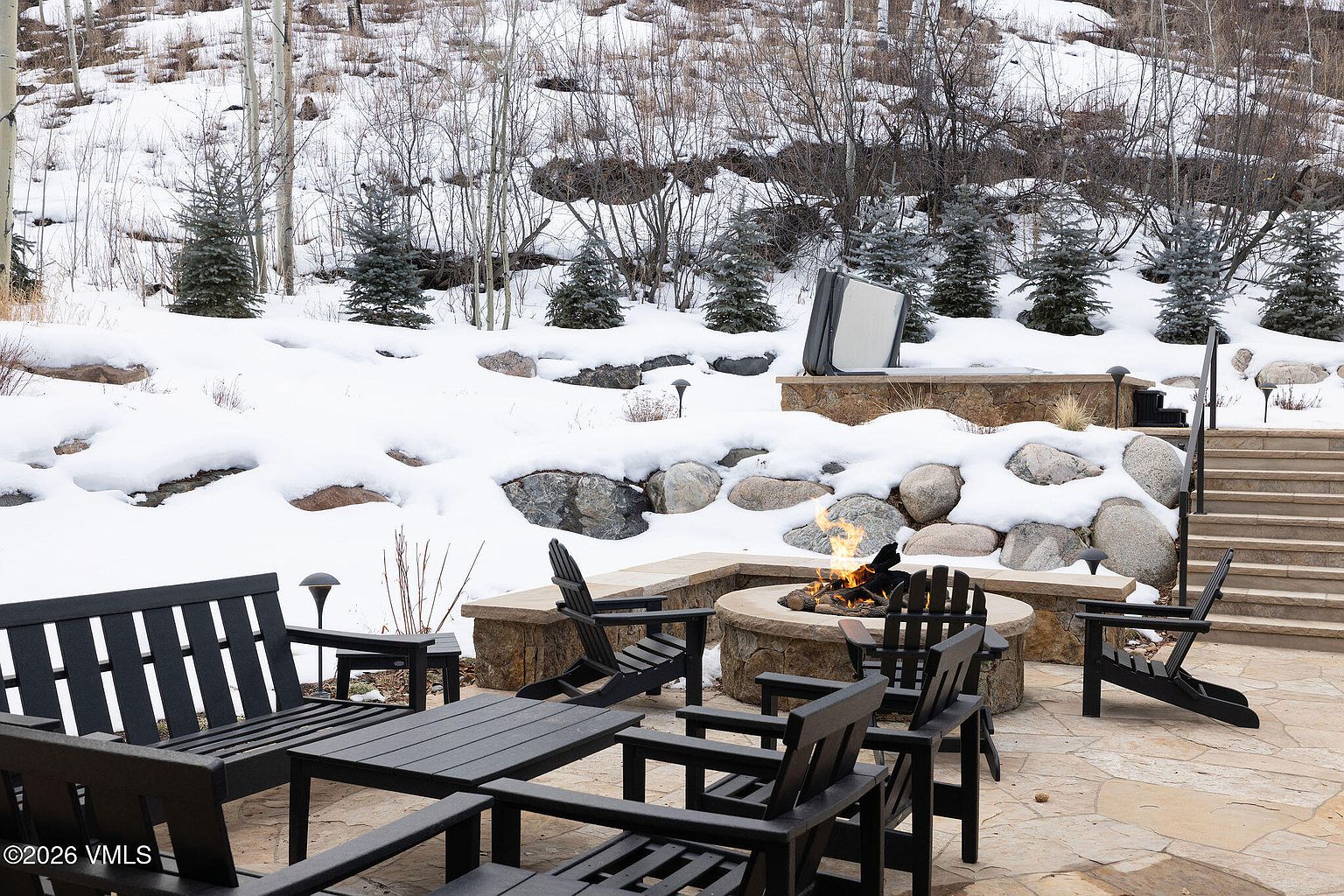 This image showcases an inviting outdoor patio area, complete with a stone fire pit surrounded by comfortable black Adirondack chairs and a matching table and bench. The patio is paved with flagstone and is nestled against a snowy hillside with mature trees and landscaping. A hot tub is visible in the background, suggesting a luxurious and relaxing outdoor living space.