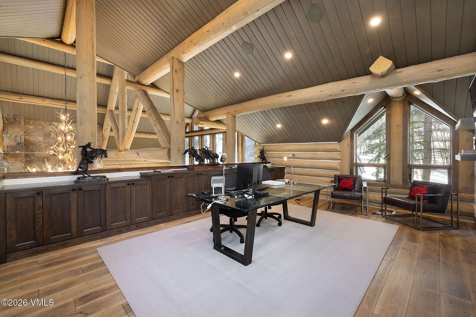 This is an interior shot of a rustic office space featuring exposed wooden beams and a log cabin-style wall. A glass-topped desk with a black frame sits on a light-colored rug, and two leather chairs with decorative pillows are positioned near a window. The room is well-lit with recessed lighting and natural light, creating a warm and inviting atmosphere.