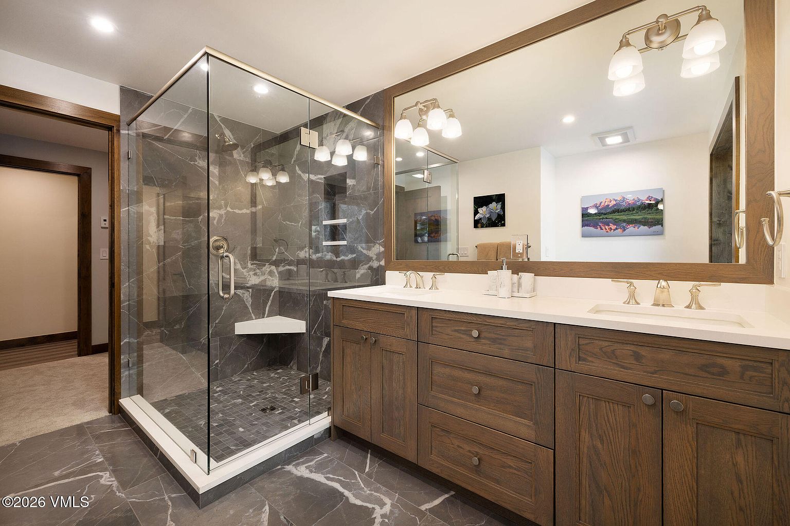 This is a primary bathroom featuring a glass-enclosed shower with gray marble-like tile and a built-in bench. The vanity has dark wood cabinets, a white countertop with double sinks, and a large mirror with sconce lighting. The floor is also a gray marble-like tile, creating a cohesive and luxurious aesthetic.