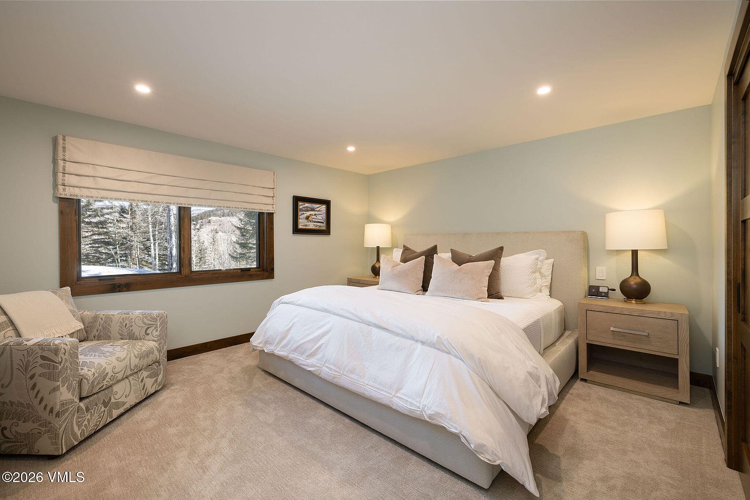 This is a well-lit primary bedroom featuring a king-size bed with white linens and neutral-toned pillows. The room has a cozy armchair near a window with a view of snow-covered trees, and a wooden nightstand with a lamp on either side of the bed. The walls are painted in a soft, muted green, creating a serene and inviting atmosphere.