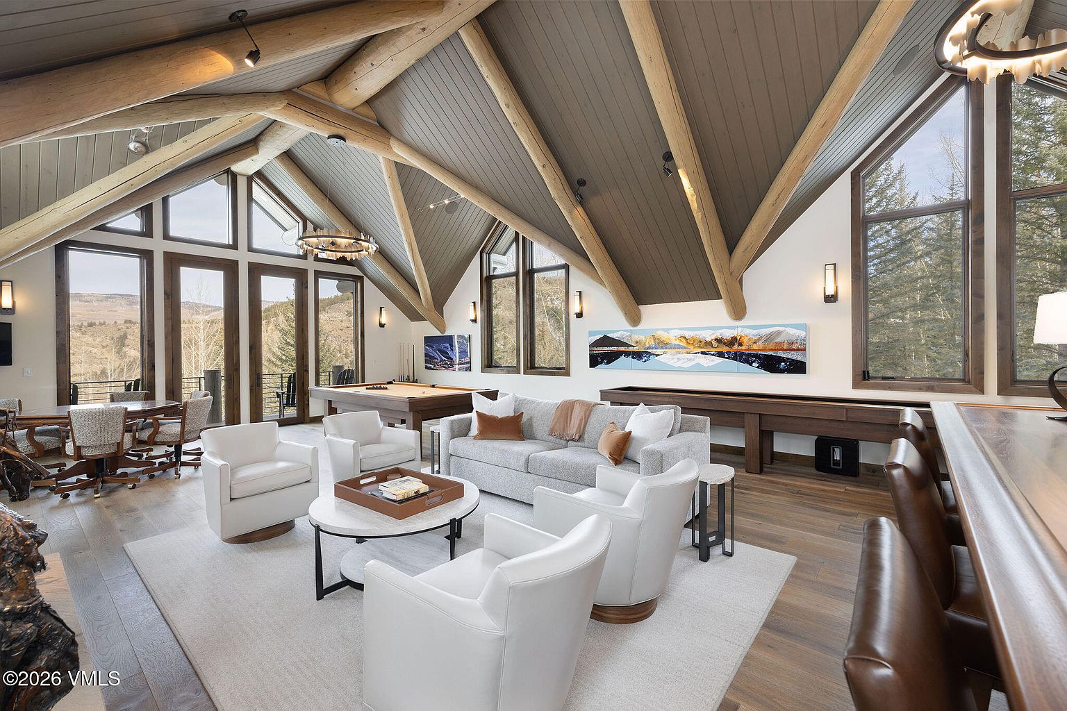 This is an interior shot of a spacious living room featuring a high vaulted ceiling with exposed wooden beams. The room is furnished with a light gray sofa, white armchairs, and a round coffee table, all arranged on a large area rug. Natural light floods the space through large windows, offering views of the surrounding landscape, and a pool table and shuffleboard table add to the entertainment options.