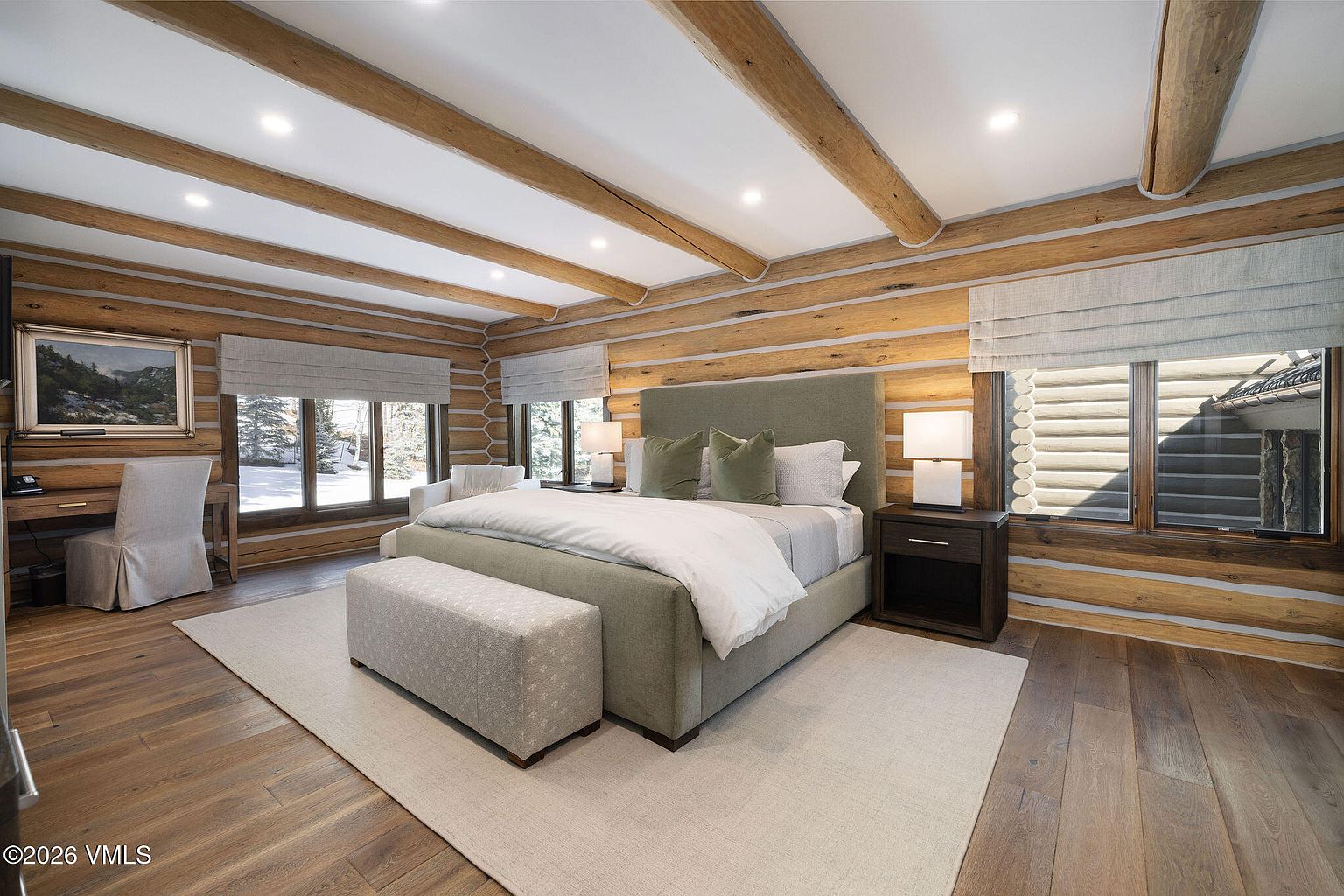 This is a primary bedroom featuring a log cabin style with exposed wooden beams and walls. The room is furnished with a large bed, a bench, and a nightstand. Natural light streams in through the windows, complemented by recessed lighting, creating a warm and inviting atmosphere.
