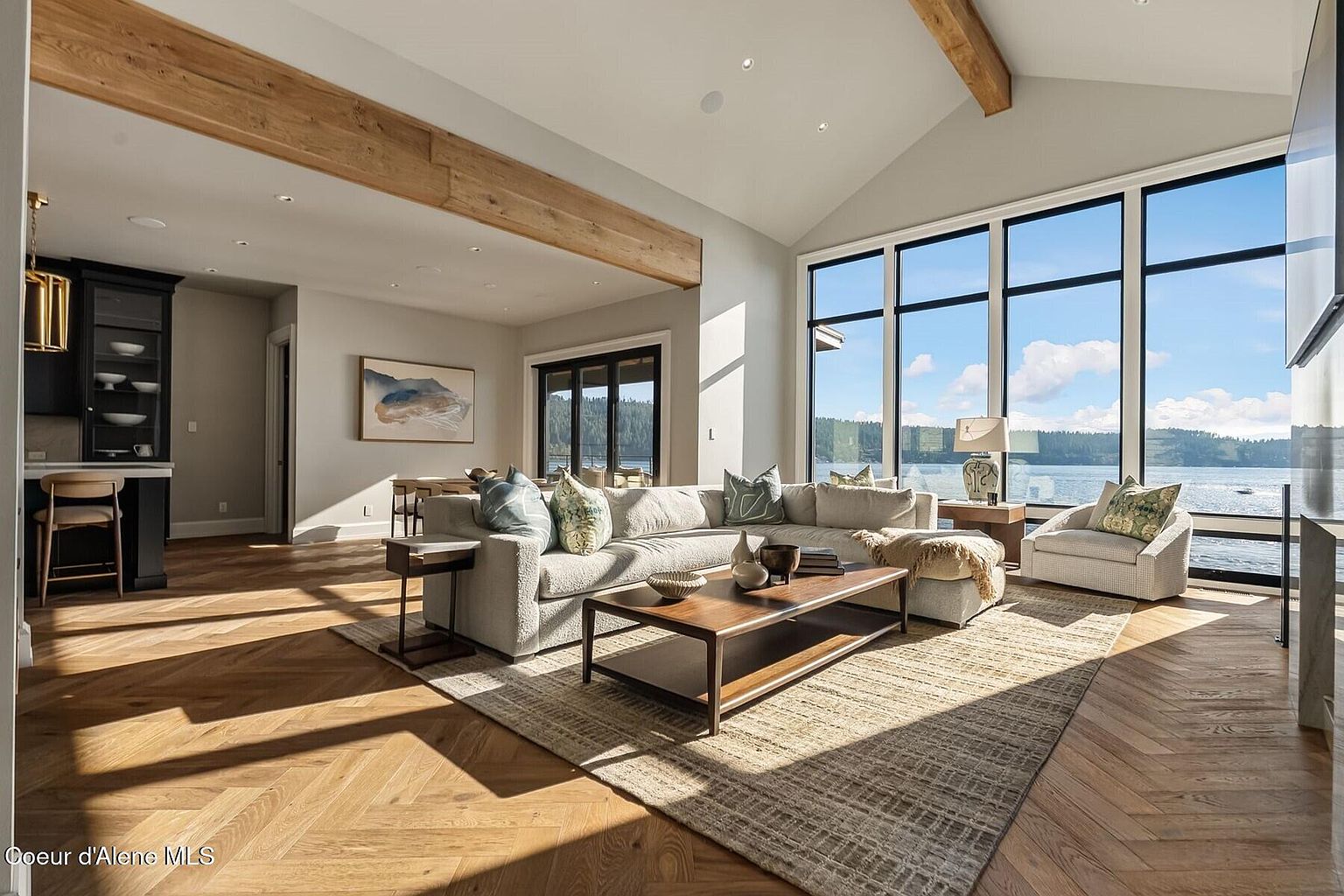 This is an interior shot of a luxurious living room featuring a large sectional sofa with decorative pillows, a wooden coffee table, and a neutral-toned area rug. The room is bathed in natural light from expansive windows offering a view of the water, and exposed wooden beams add a rustic touch to the modern design. The herringbone wood flooring adds a touch of elegance.