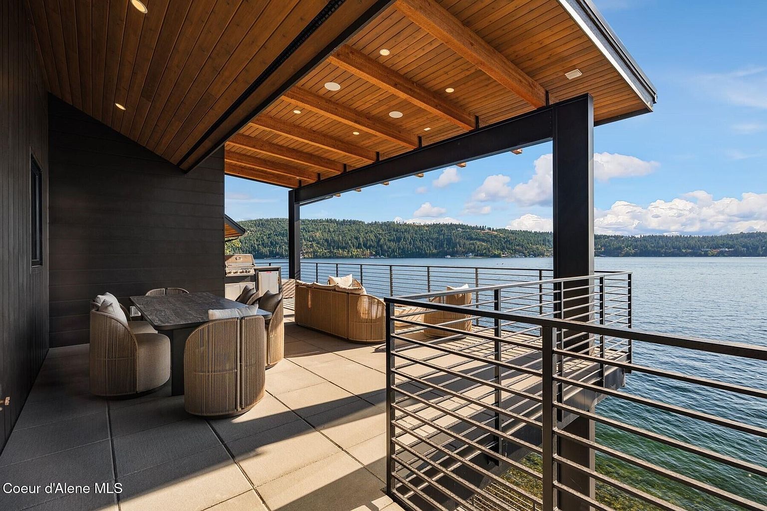 This image showcases a luxurious outdoor patio/deck area with stunning waterfront views. The space is furnished with a dining table and chairs, as well as a comfortable seating area, perfect for relaxation and entertaining. The modern design features a wooden ceiling, sleek metal railings, and a neutral color palette, creating an inviting and sophisticated atmosphere.