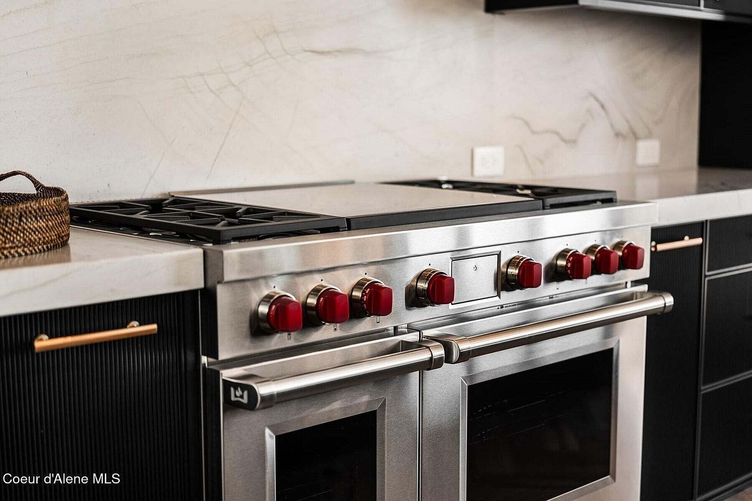 This image showcases a high-end kitchen featuring a stainless steel range with red knobs, set against a backdrop of marble backsplash and countertops. The cabinetry is a dark, textured material with sleek, gold-toned hardware, contributing to a modern and luxurious aesthetic. The focus is on the appliance, highlighting its professional-grade quality and design.