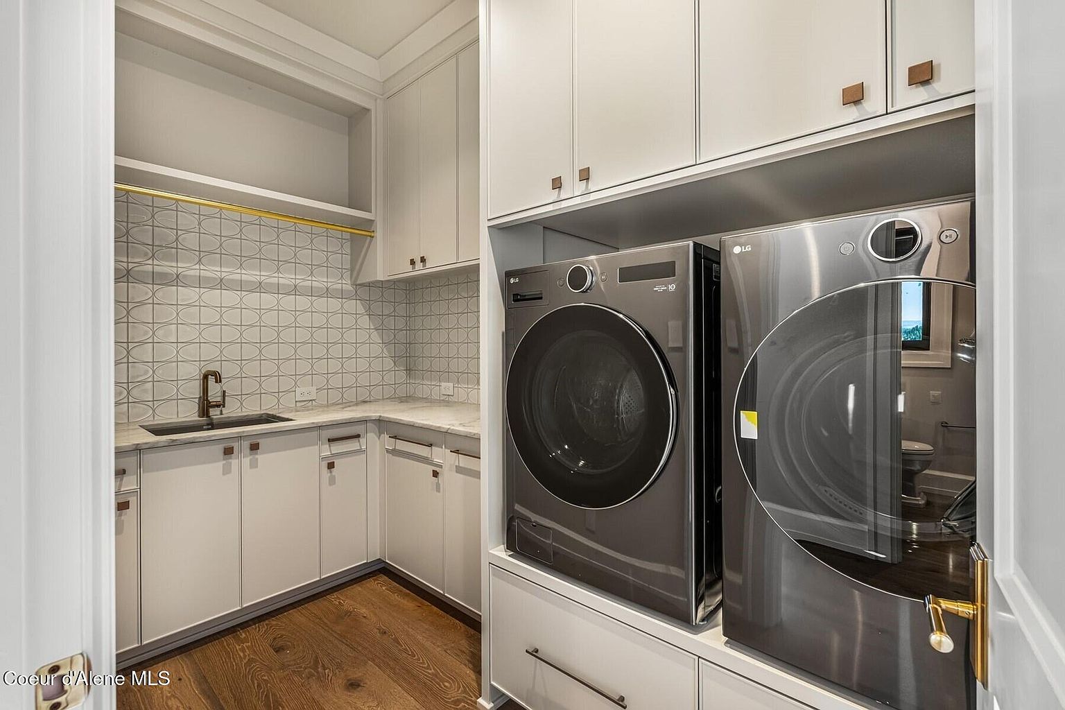 This is a well-organized laundry room featuring modern LG washer and dryer units stacked on a white base cabinet with drawers. The room includes white cabinetry with bronze hardware, a sink area with a patterned backsplash, and hardwood flooring. The overall impression is clean, efficient, and stylish.