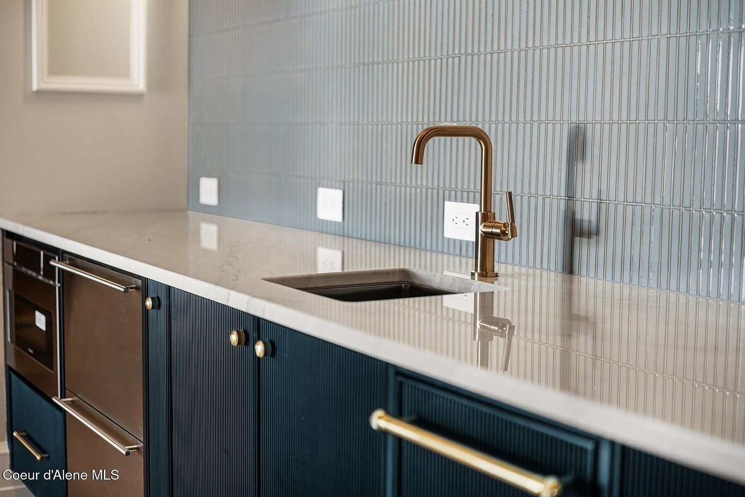 This image showcases a modern kitchen detail, focusing on the sink area with a stylish gold faucet and a dark sink. The backsplash features vertically stacked, light blue tiles, complemented by a light-colored countertop. Below the counter, dark blue cabinets with gold hardware add a touch of elegance.
